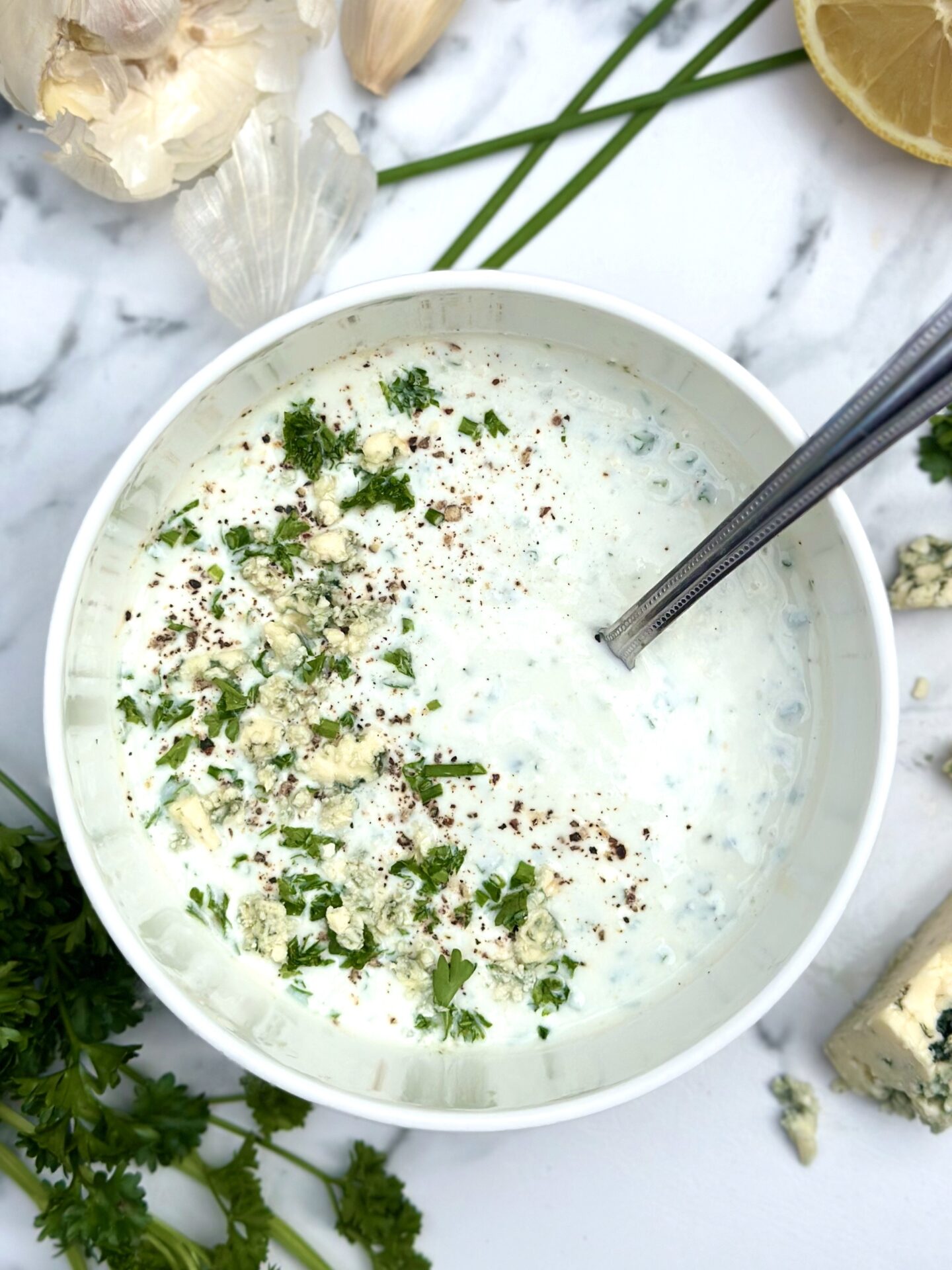 Bowl of homemade blue cheese dressing seen on a white marble countertop surrounded by lemon, garlic and fresh herbs