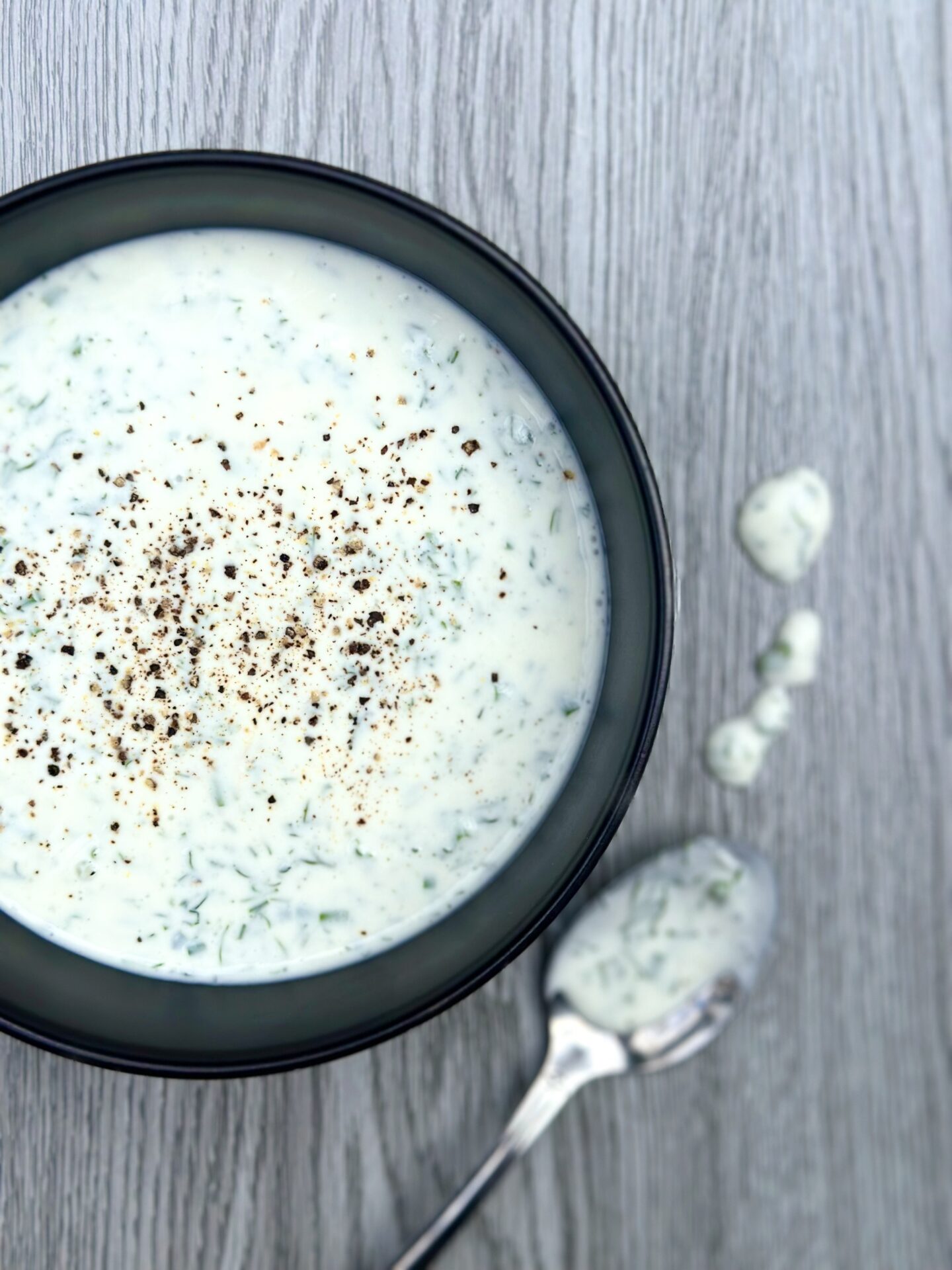 A bowl of creamy and fresh homemade buttermilk ranch dressing, with a spoon and drips of the dressing on a grey wood board