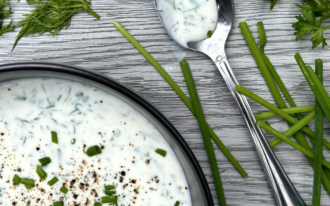 A bowl of fresh buttermilk ranch dressing on a grey wood board, surrounded by fresh chives, dill and parsley