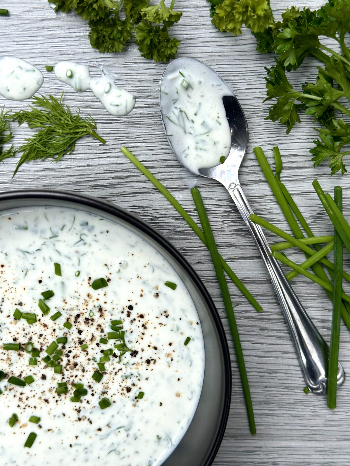 A bowl of fresh buttermilk ranch dressing on a grey wood board, surrounded by fresh chives, dill and parsley