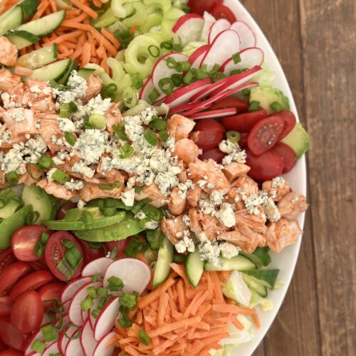 A large platter of Buffalo Chicken Dinner Salad full of bright fresh vegetables and big chunks of buffalo chicken is seen from above on a wood table