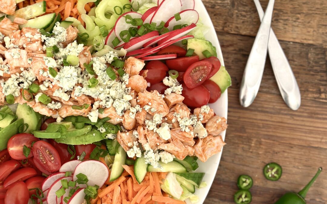 A large platter of Buffalo Chicken Dinner Salad full of bright and crisp vegetables is seen from above on a wood table with serving spoons and sliced jalapeños