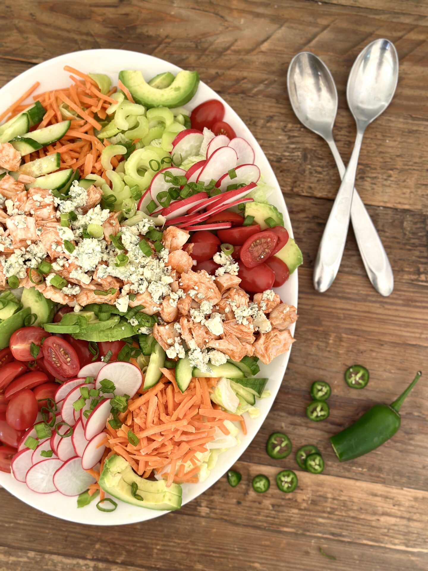 A large platter of Buffalo Chicken Dinner Salad full of bright and crisp vegetables is seen from above on a wood table with serving spoons and sliced jalapeños