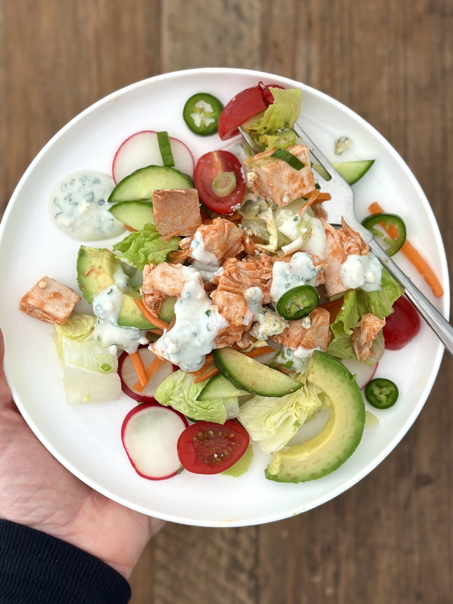 A hand holds a plate of Buffalo Chicken Dinner Salad full of fresh vegetables and chucks of Buffalo Chicken