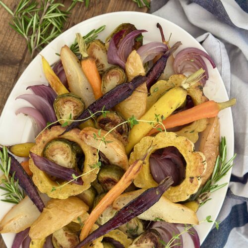 A platter of Maple Butter Roasted Vegetables with Pears is seen from above on a wood table surrounded by fresh rosemary and a grey striped kitchen cloth.