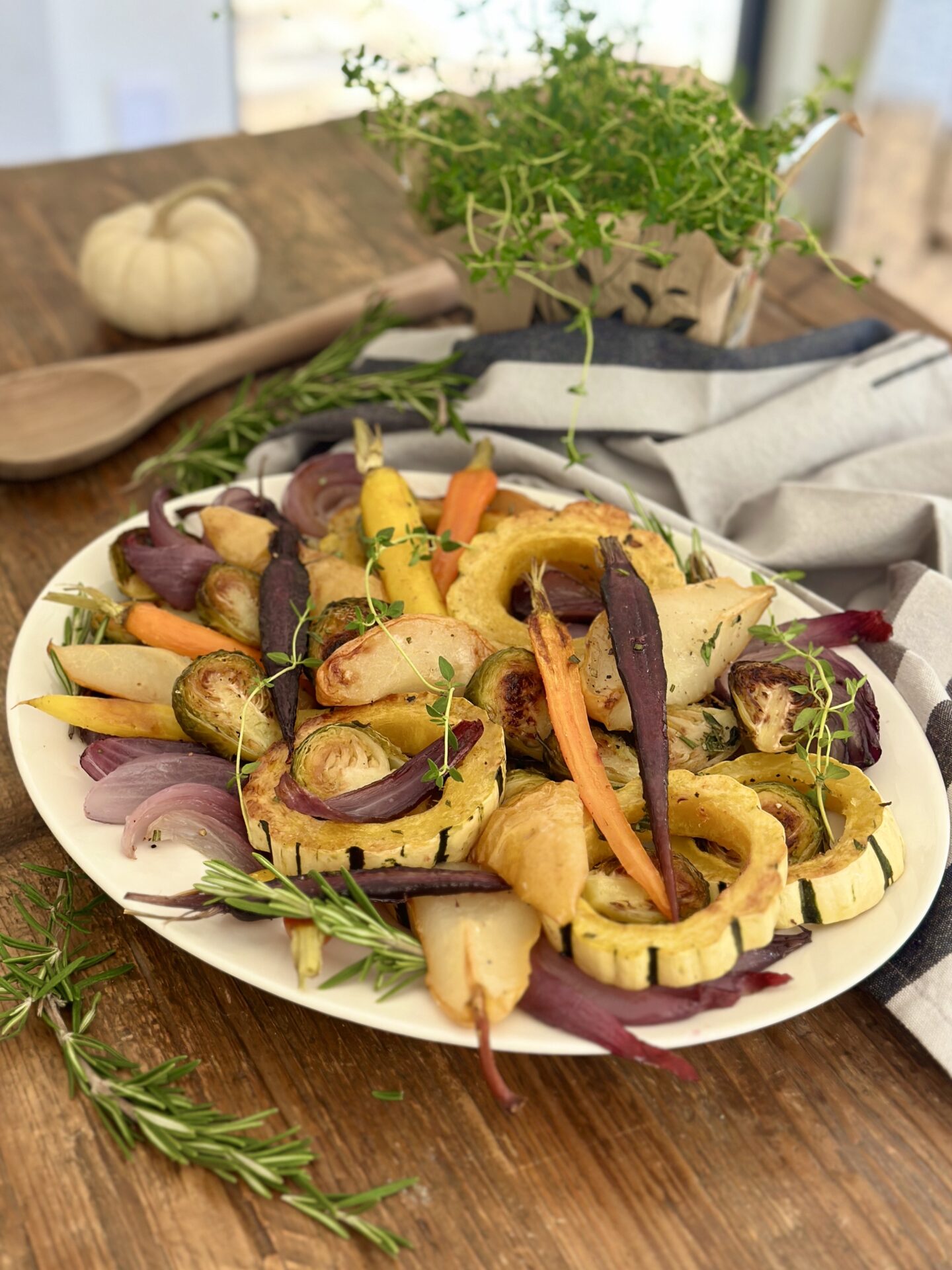 A Thanksgiving side dish of Maple Butter Roasted Vegetables with Pears sits on a wood table, surrounded by fresh herbs