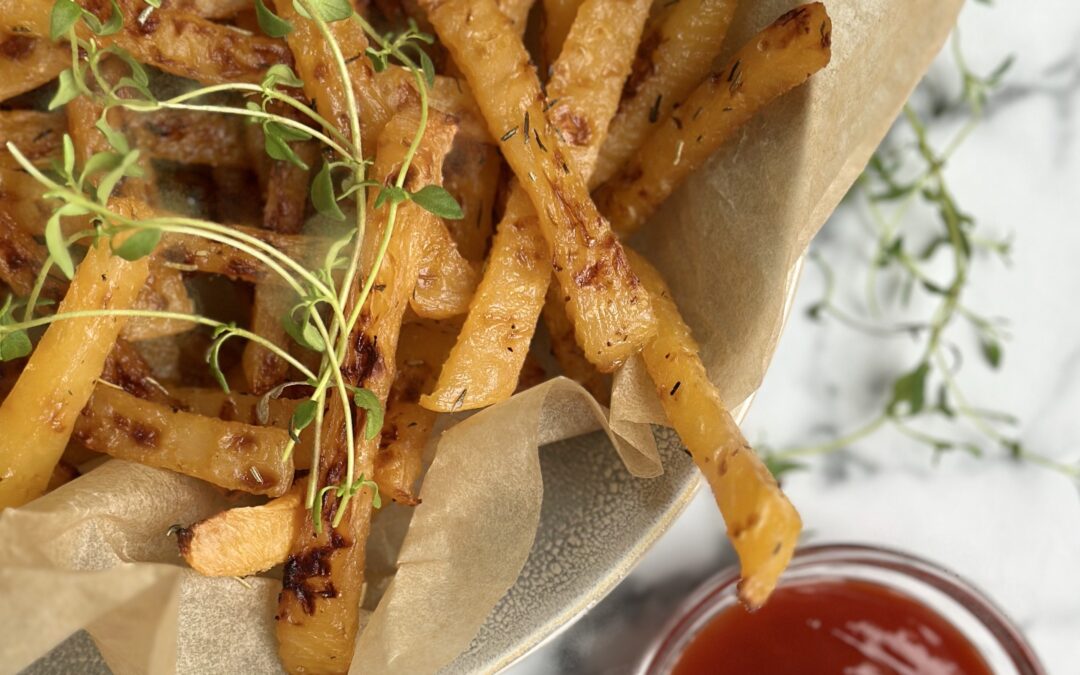 A brown paper lined bowl of golden rutabaga fries garnished with fresh thyme sits on a white marble counter top along with a small bowl of red ketchup