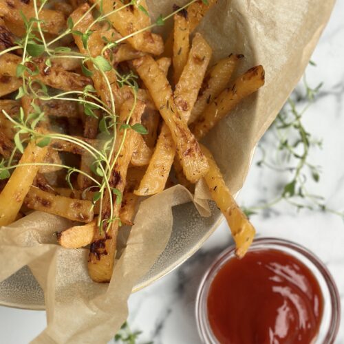 A brown paper lined bowl of golden rutabaga fries garnished with fresh thyme sits on a white marble counter top along with a small bowl of red ketchup