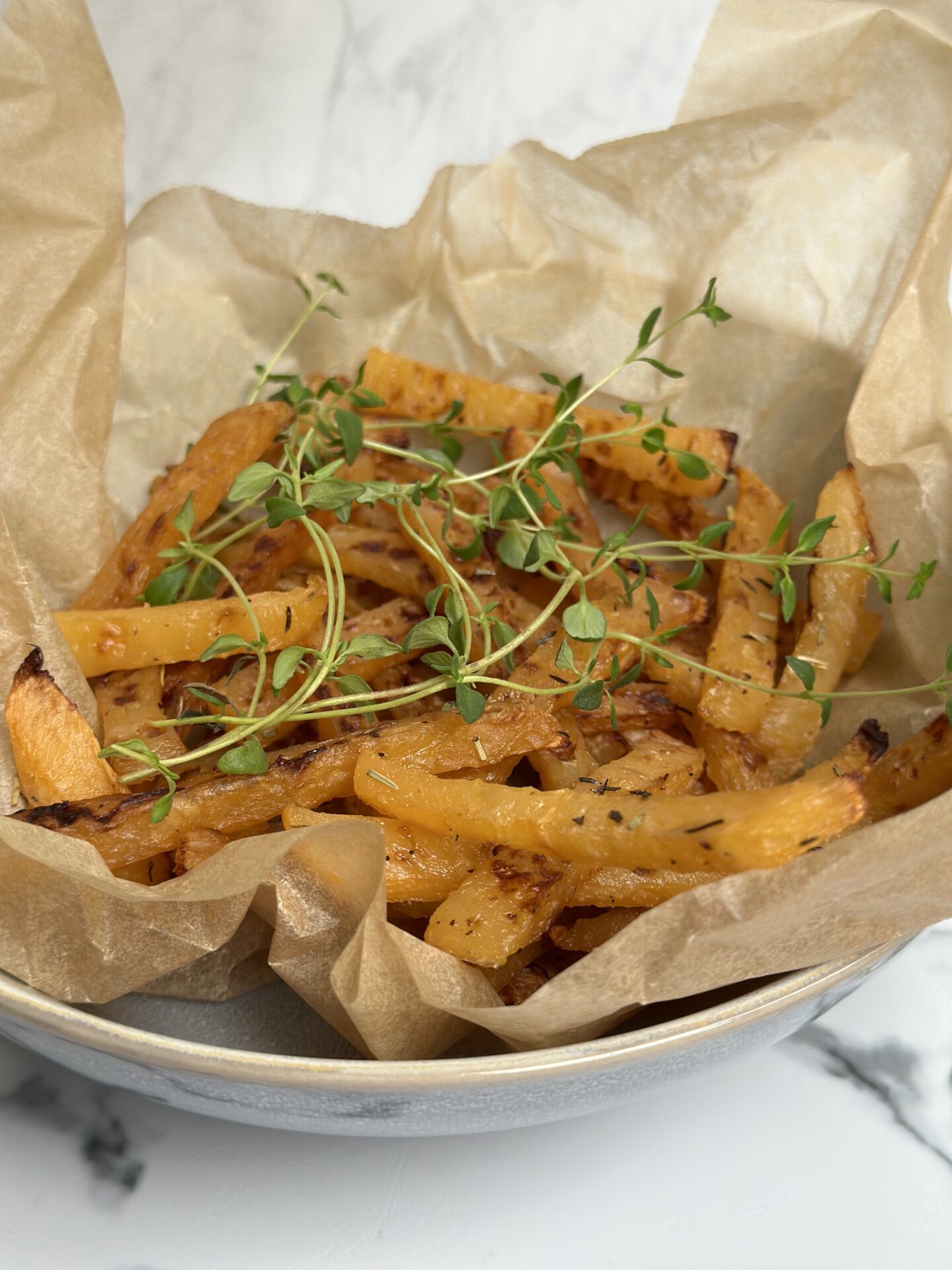 A brown paper lined bowl of golden oven roasted rutabaga fries sits on a white marble counter top