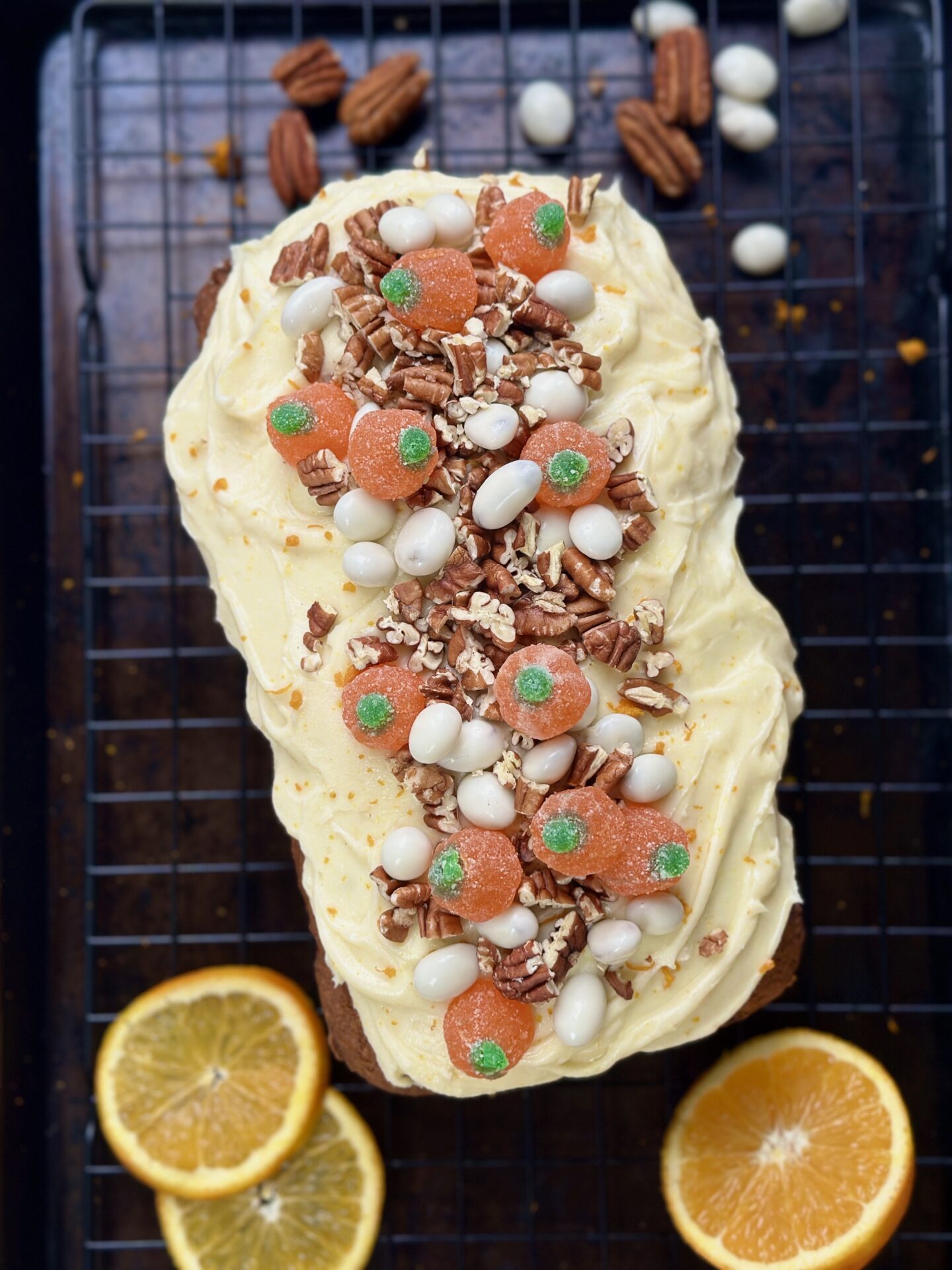 A decorated loaf with swirls of thick orange cream cheese frosting and loads of candy and pecan toppings is seen from above on a black baking sheet