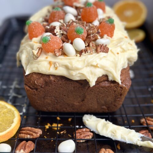 Pumpkin Bread with Orange Cream Cheese Frosting is seen on a cooling rack, surrounded by the frosting knife, pecans and orange slices