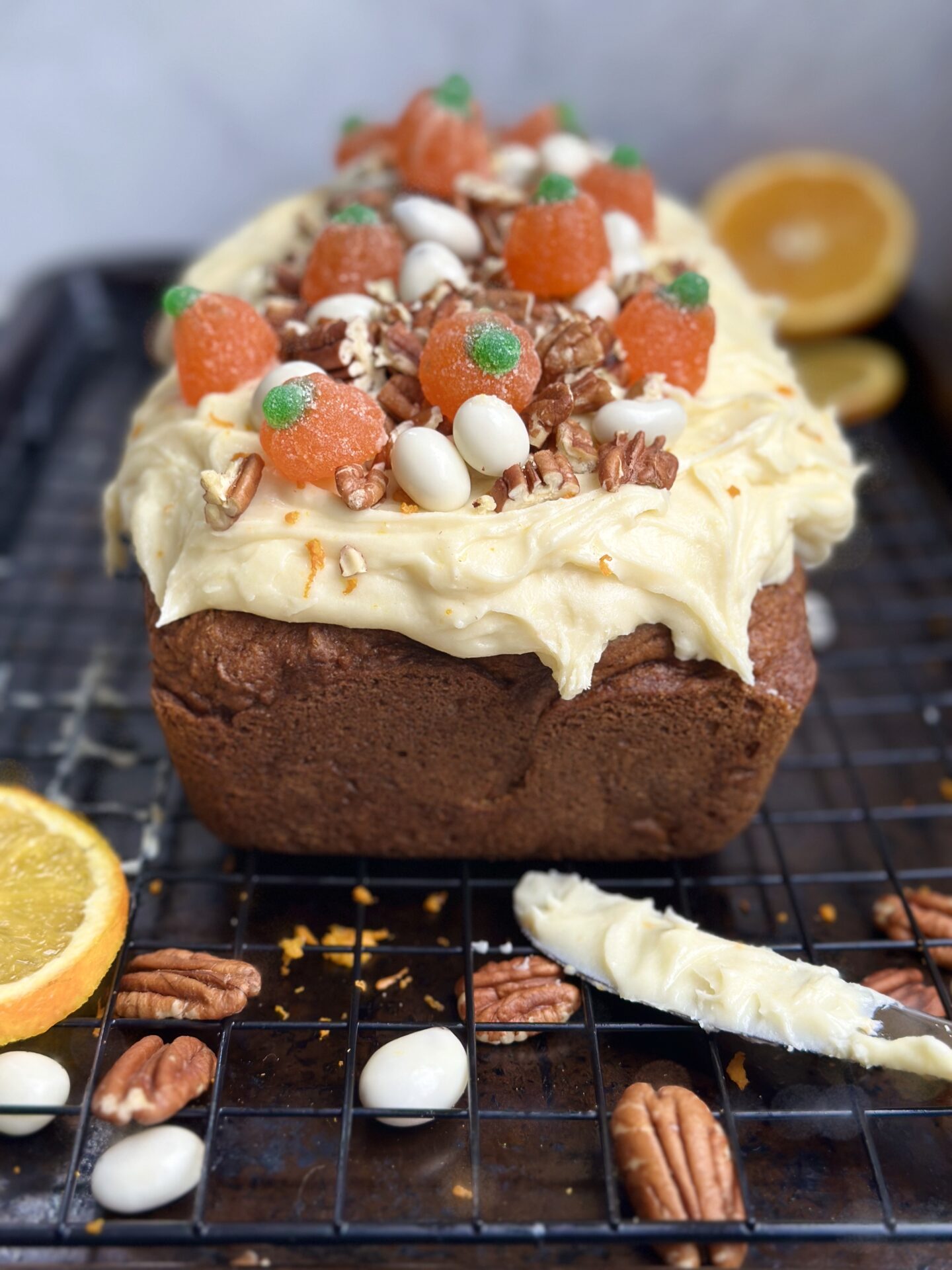 Pumpkin Bread with Orange Cream Cheese Frosting is seen on a cooling rack, surrounded by the frosting knife, pecans and orange slices