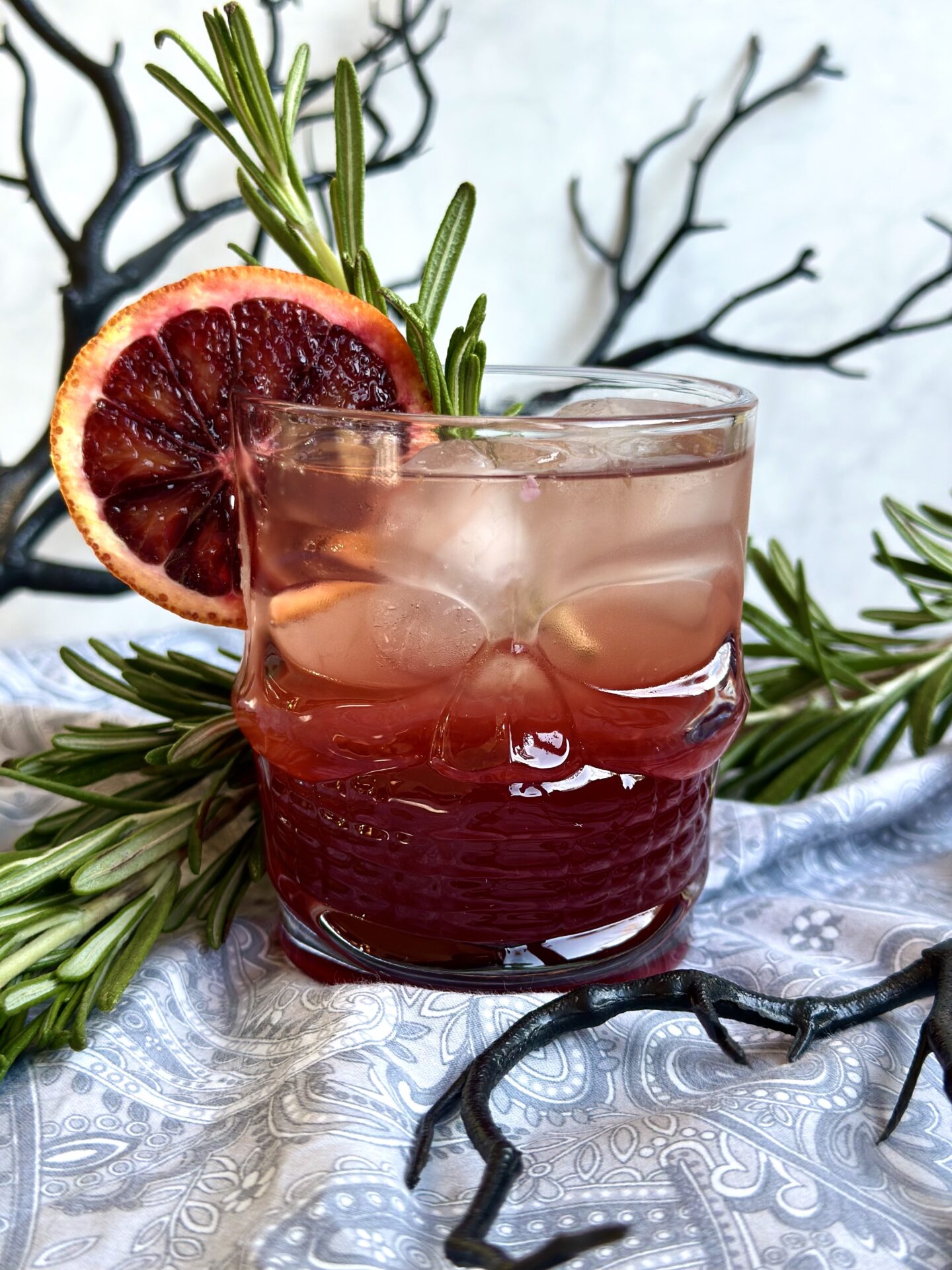 A blood orange and black currant aperol spritz is served in a halloween themed glass, surrounded by black branches