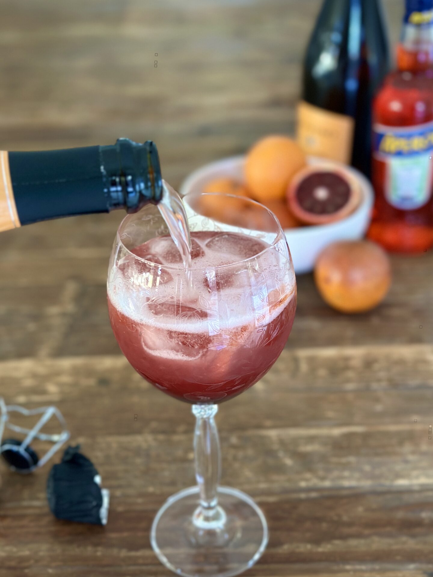 Prosecco is poured into a blood orange and black currant aperol spritz while it is prepared on a wood table