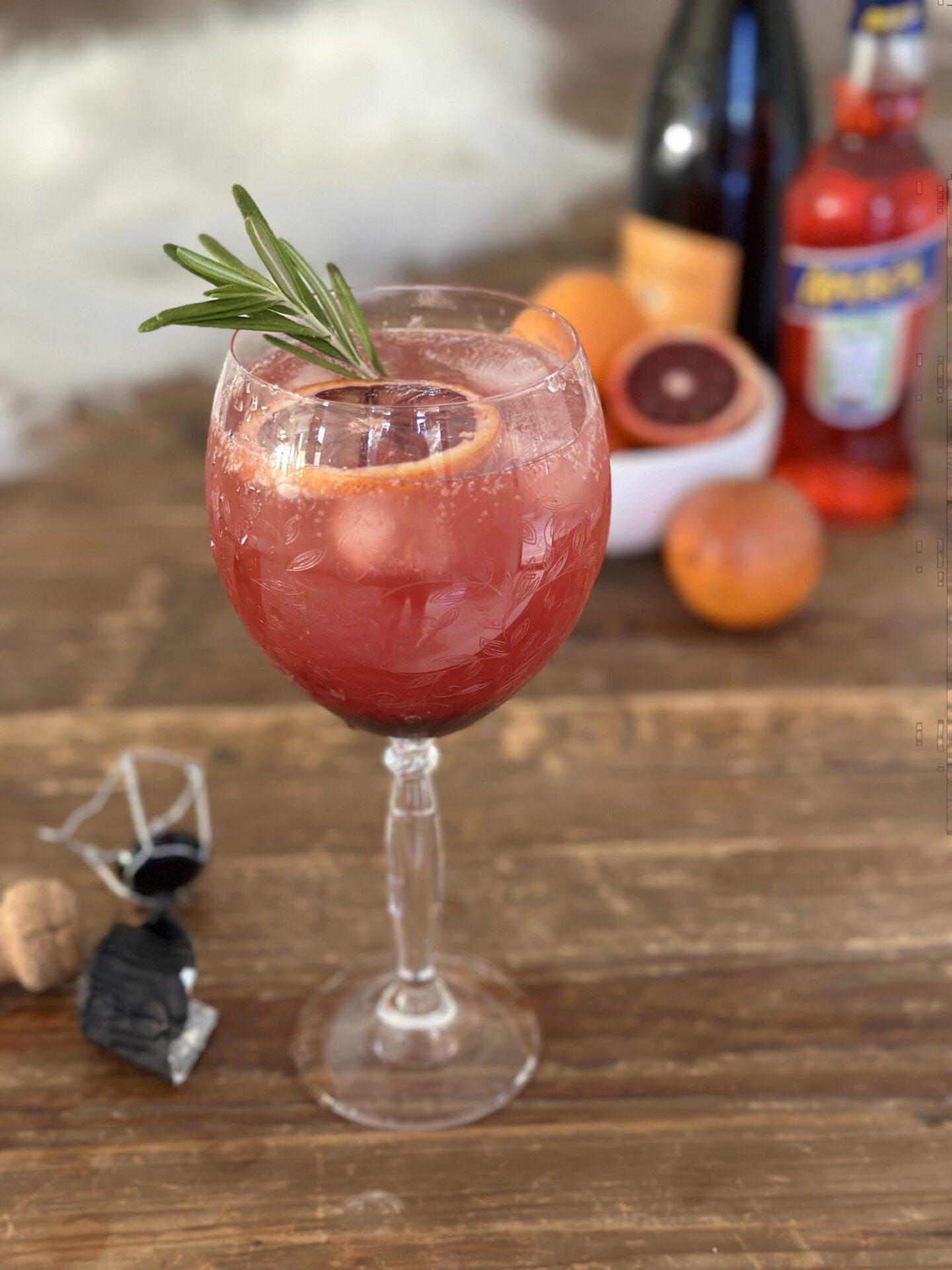 A blood orange and black currant Aperol spritz garnished with rosemary sits on a wood table