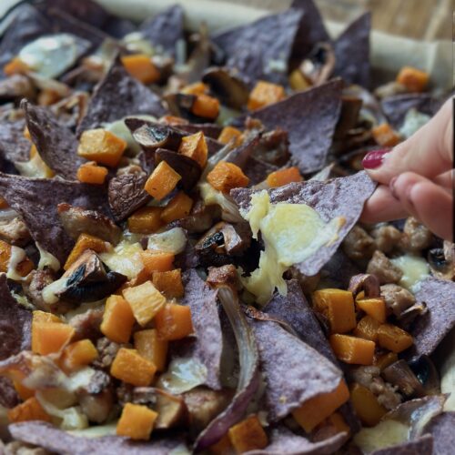 A hand lifts a cheesy chip from a platter of blue corn harvest nachos