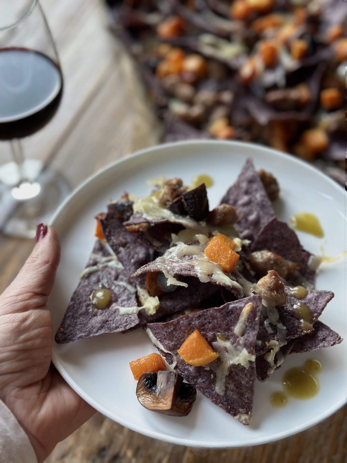 A hand holds a plate of blue corn harvest nachos in front of a glass of red wine