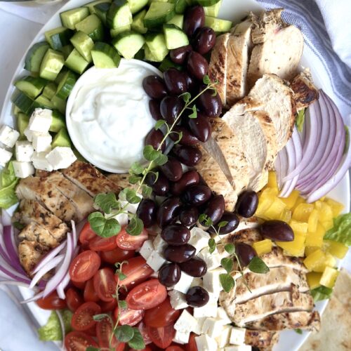 A large platter of Grilled Chicken Greek Salad is seen from above with generous amounts of tomatoes, cucumber, chicken, feta cheese and olives