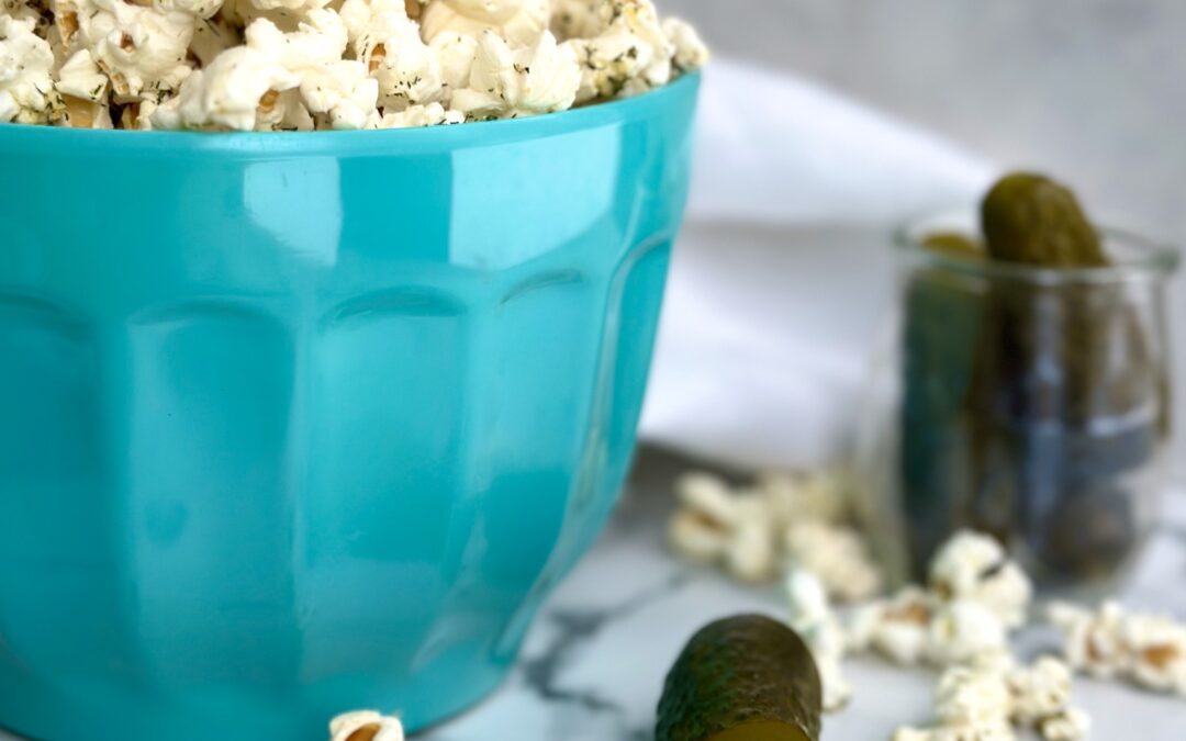 A turquoise bowl of Dill Pickle popcorn is seen on a white marble table surrounded by green dill pickles, fresh dill sprigs and popcorn pieces