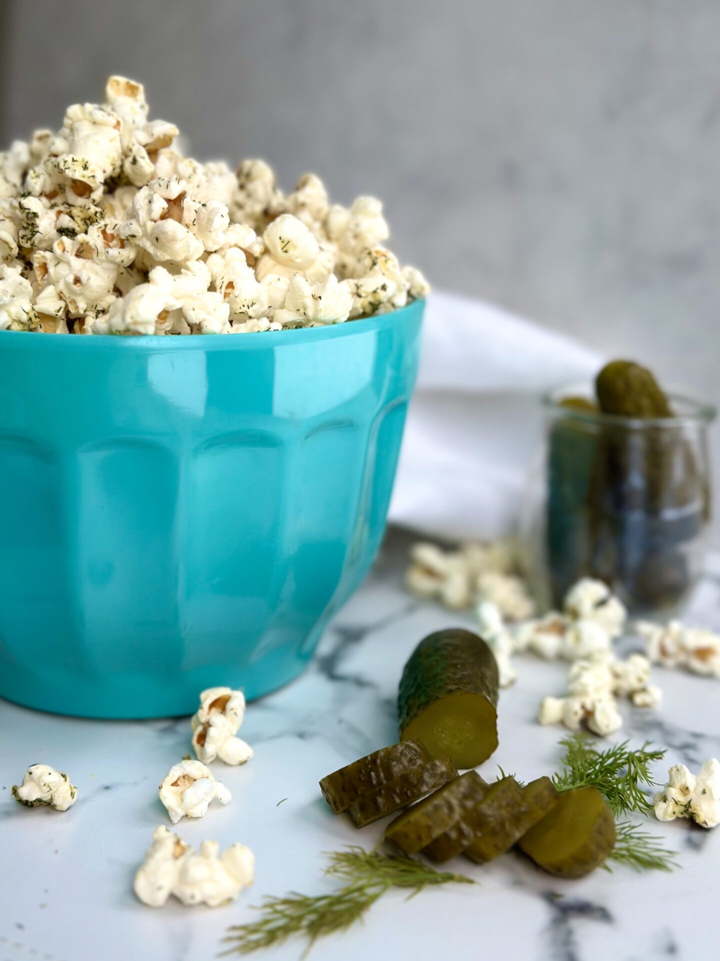 A turquoise bowl of Dill Pickle popcorn is seen on a white marble table surrounded by green dill pickles, fresh dill sprigs and popcorn pieces