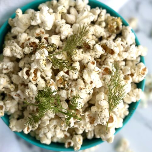 A bowl of Dill Pickle Popcorn is seen from above garnished with fresh dill sprigs and set on a white marble table