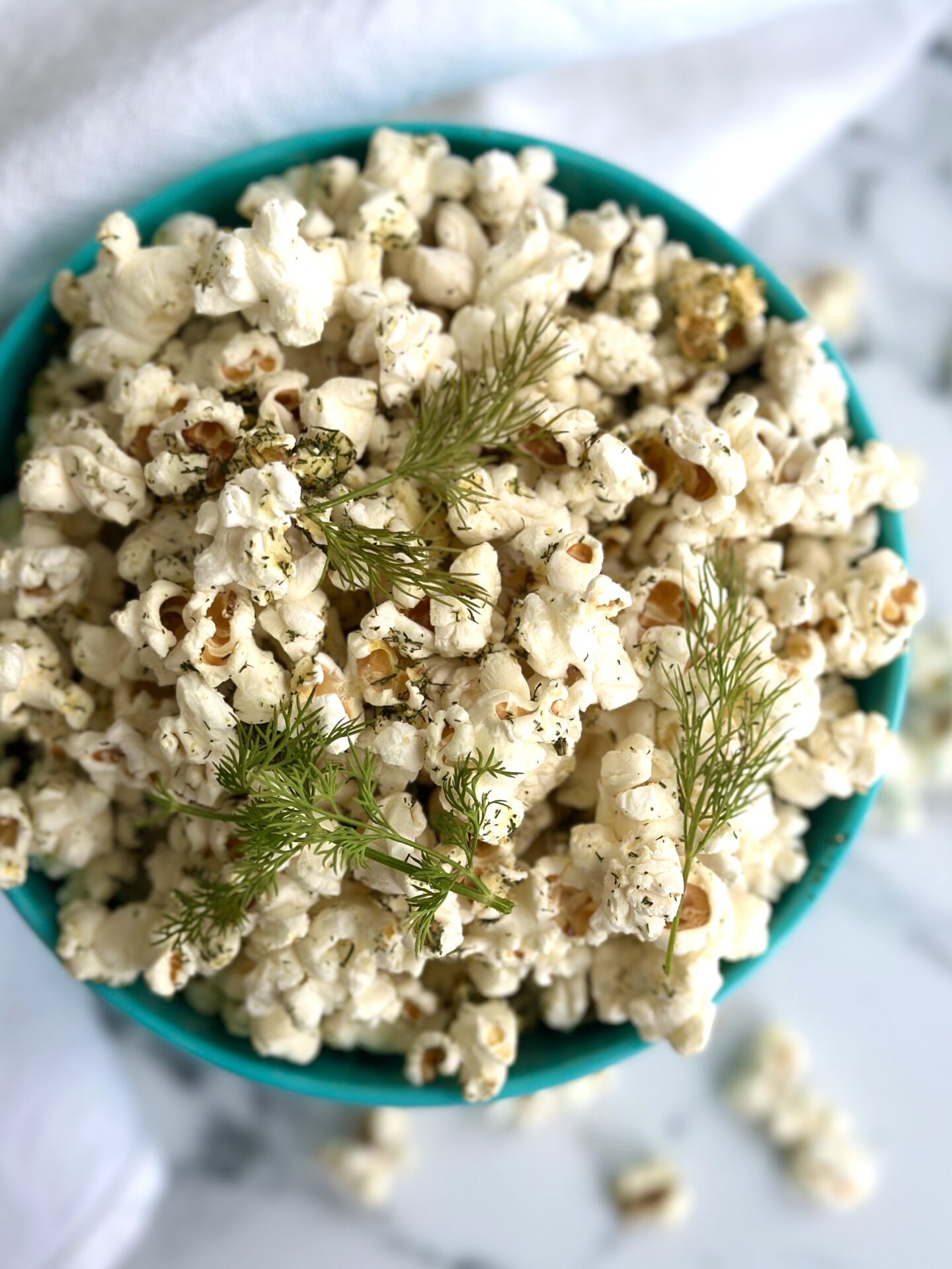 A bowl of Dill Pickle Popcorn is seen from above garnished with fresh dill sprigs and set on a white marble table
