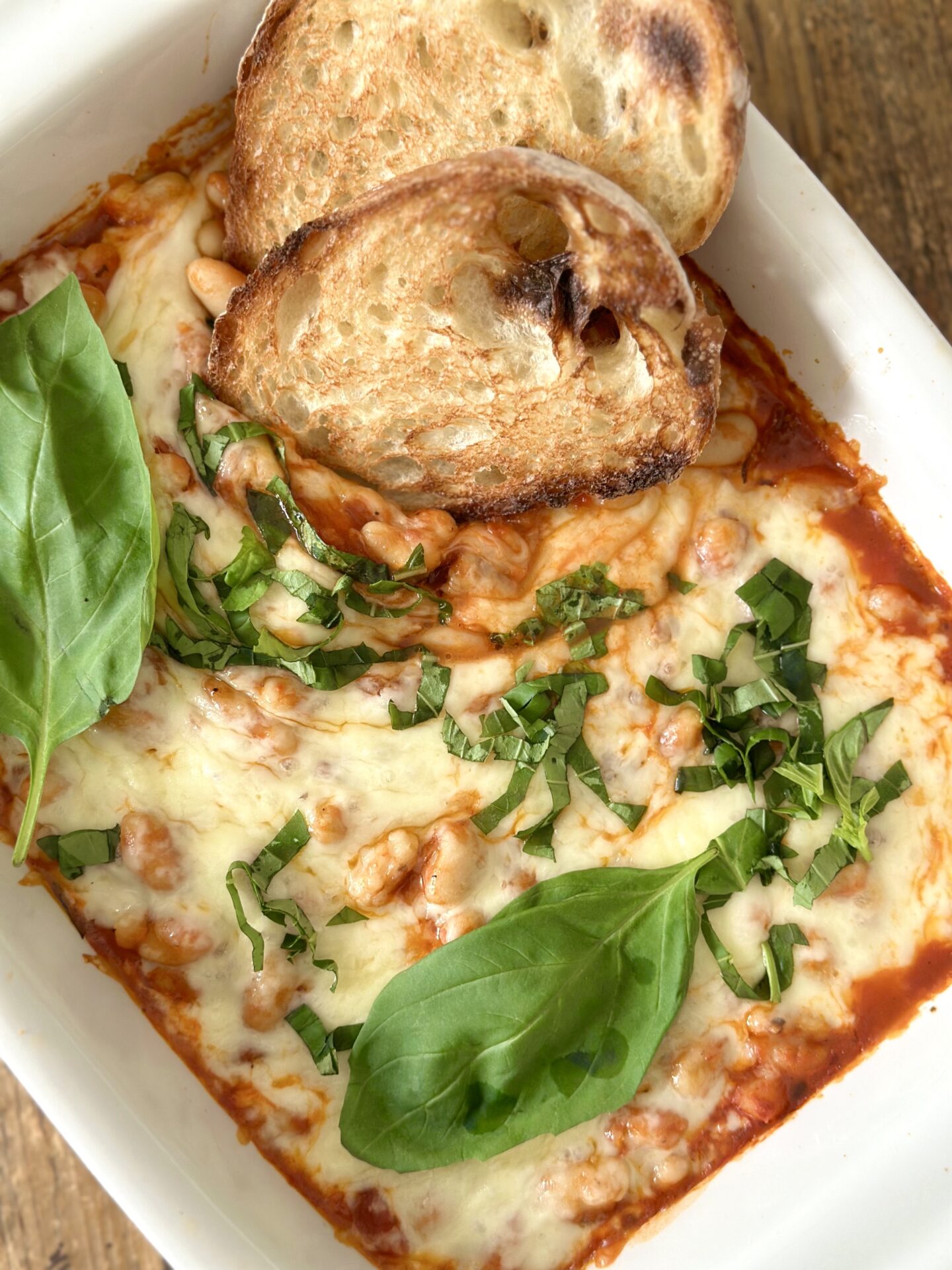 A pan of Easy Cheesy Beans is seen from above garnished with fresh basil and slices of garlic bread