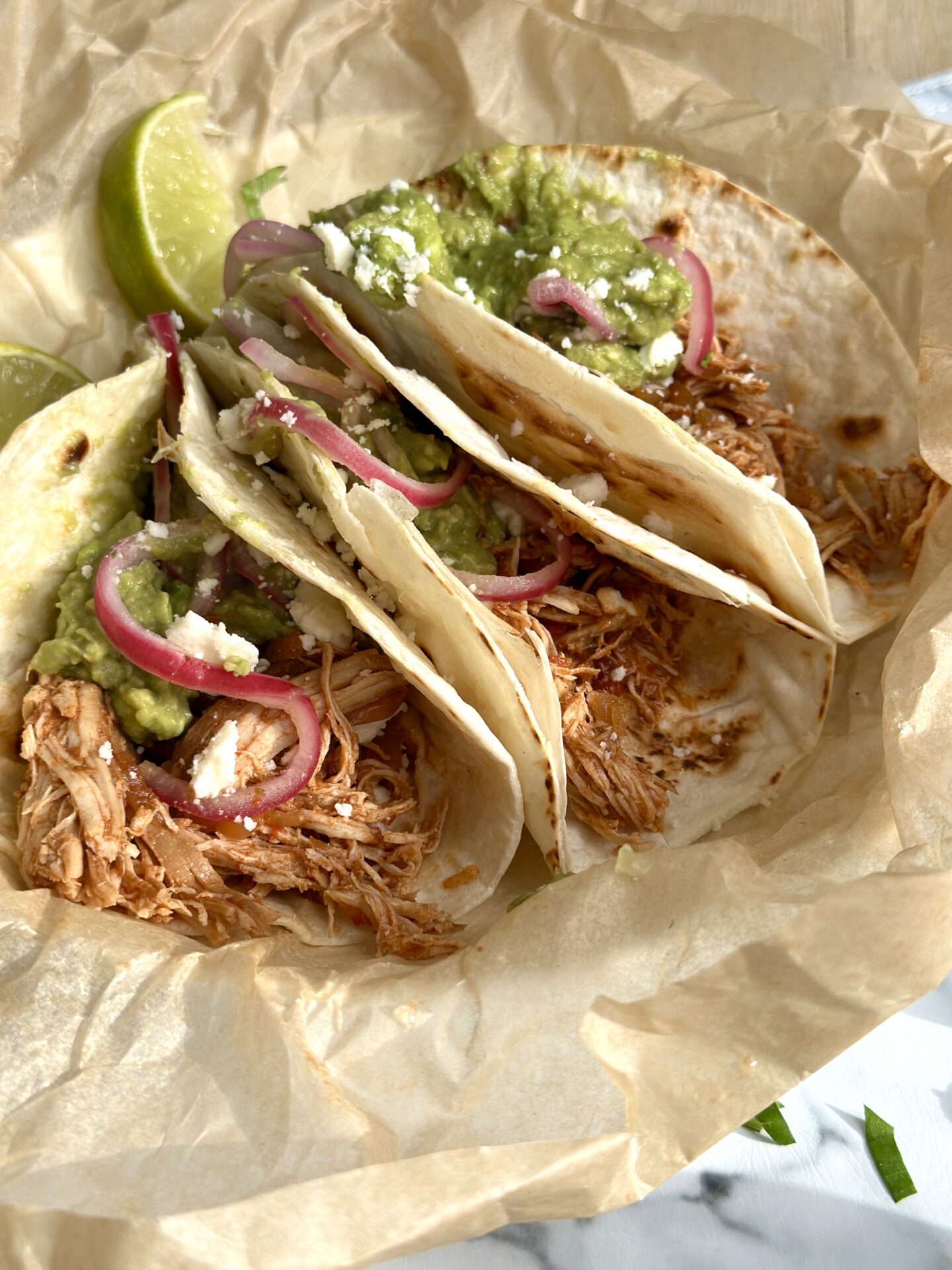 A trio of pulled chicken tacos are topped with guacamole and pickled red onions