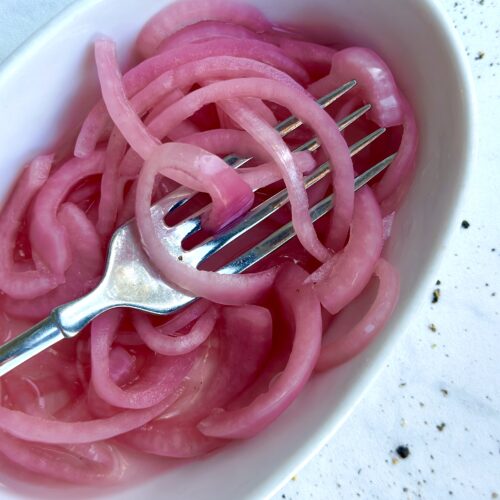 A dish of homemade bright pink pickled red onions tangled in the tines of a fork is seen from above on a white marble counter