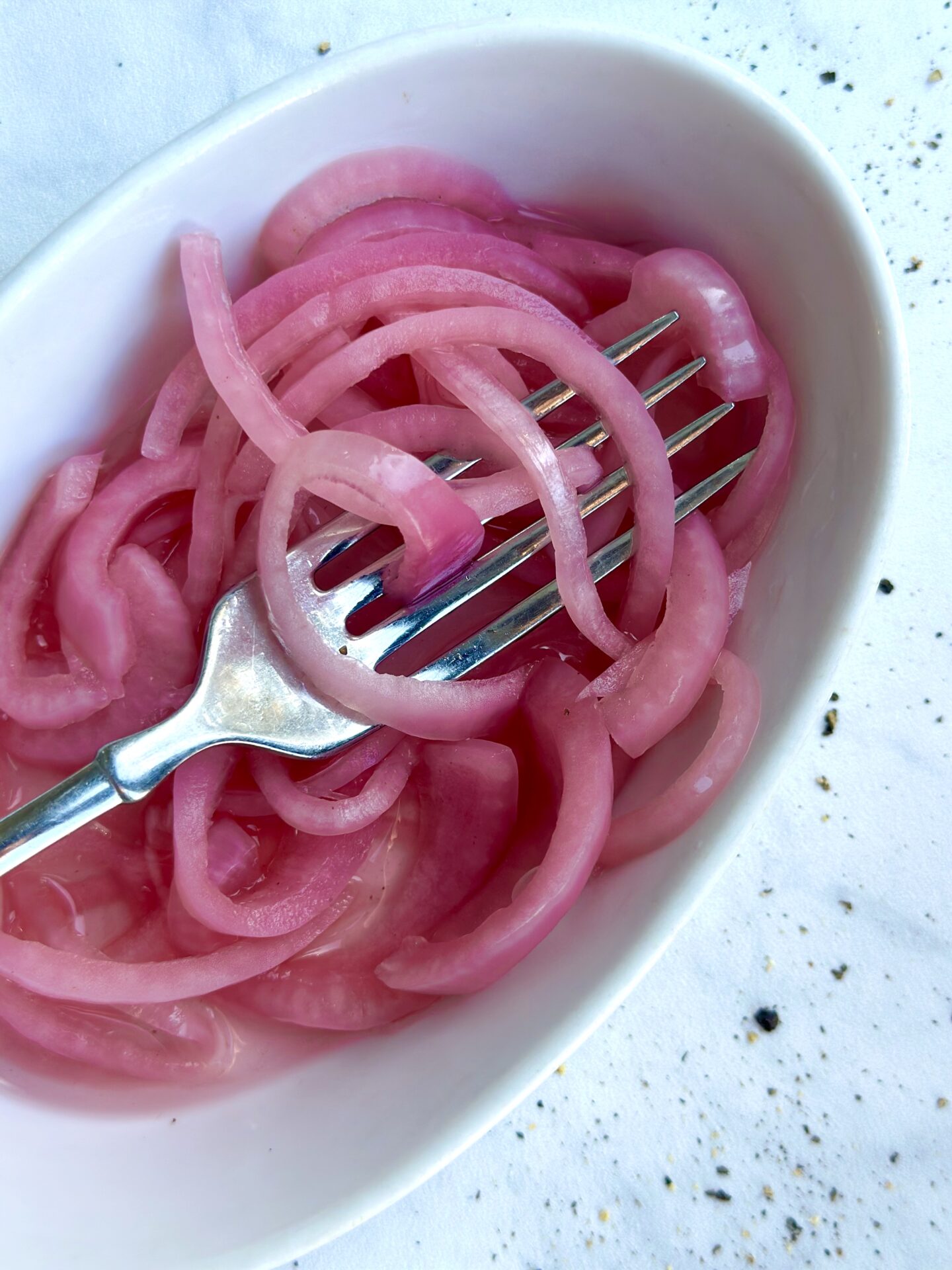 A dish of homemade bright pink pickled red onions tangled in the tines of a fork is seen from above on a white marble counter