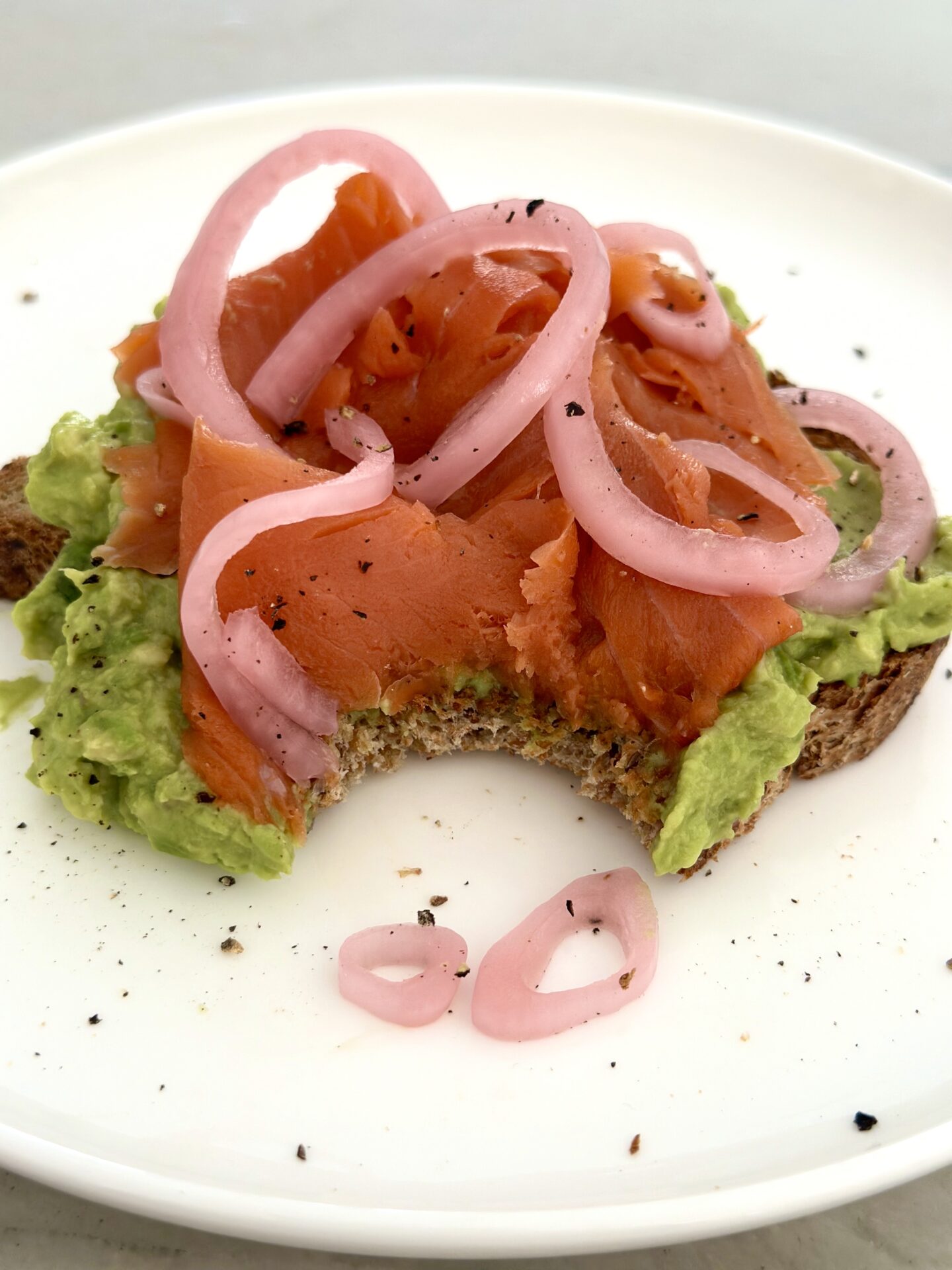 Avocado toast with smoked salmon and pickled red onions sits on a white plate.  A large bite has been taken out of the avocado toast