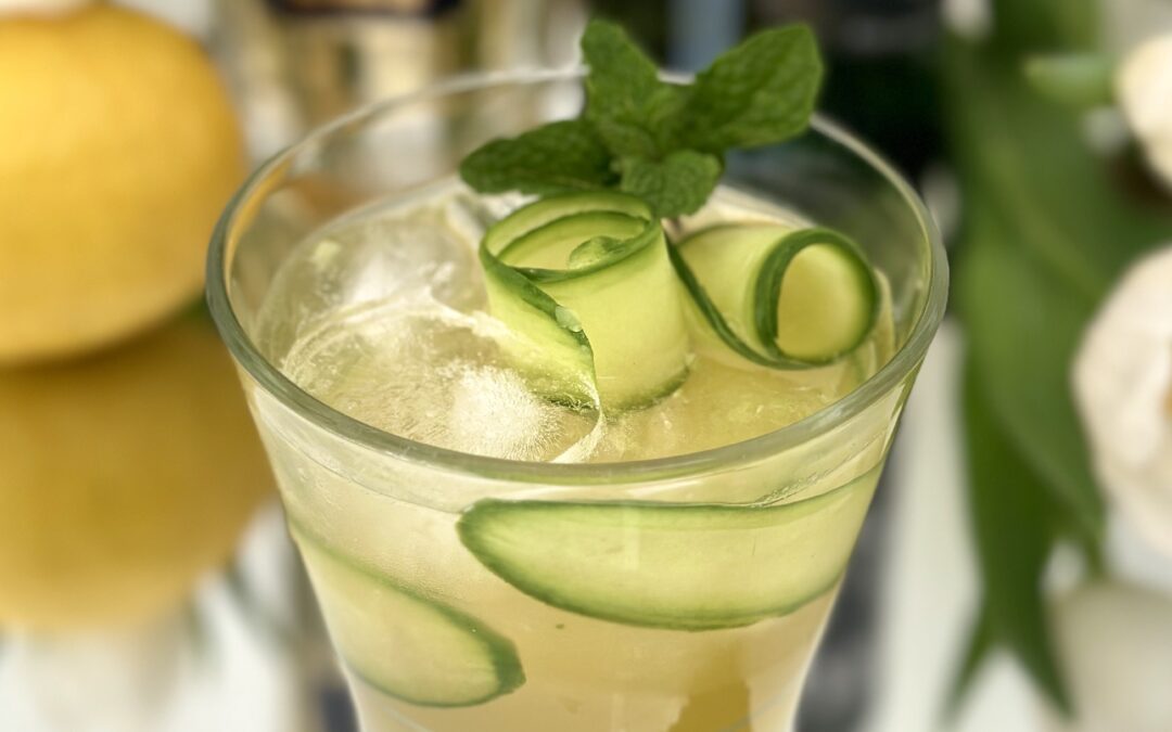 An Irish Maid Cocktail garnished with cucumber curls and fresh mint sits on a sparkling bar surrounded by bottles of Irish Whiskey and St Germain Elderflower Liqueur