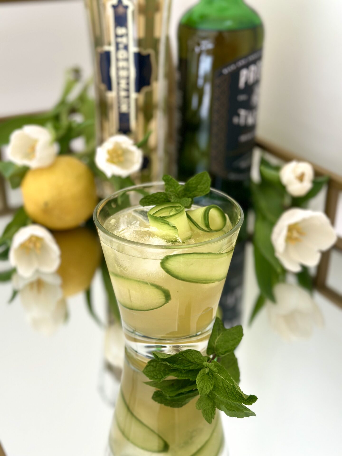 An Irish Maid Cocktail sits on a sparkling bar with Elderflower Liqueur, Irish Whiskey, lemons and white tulips