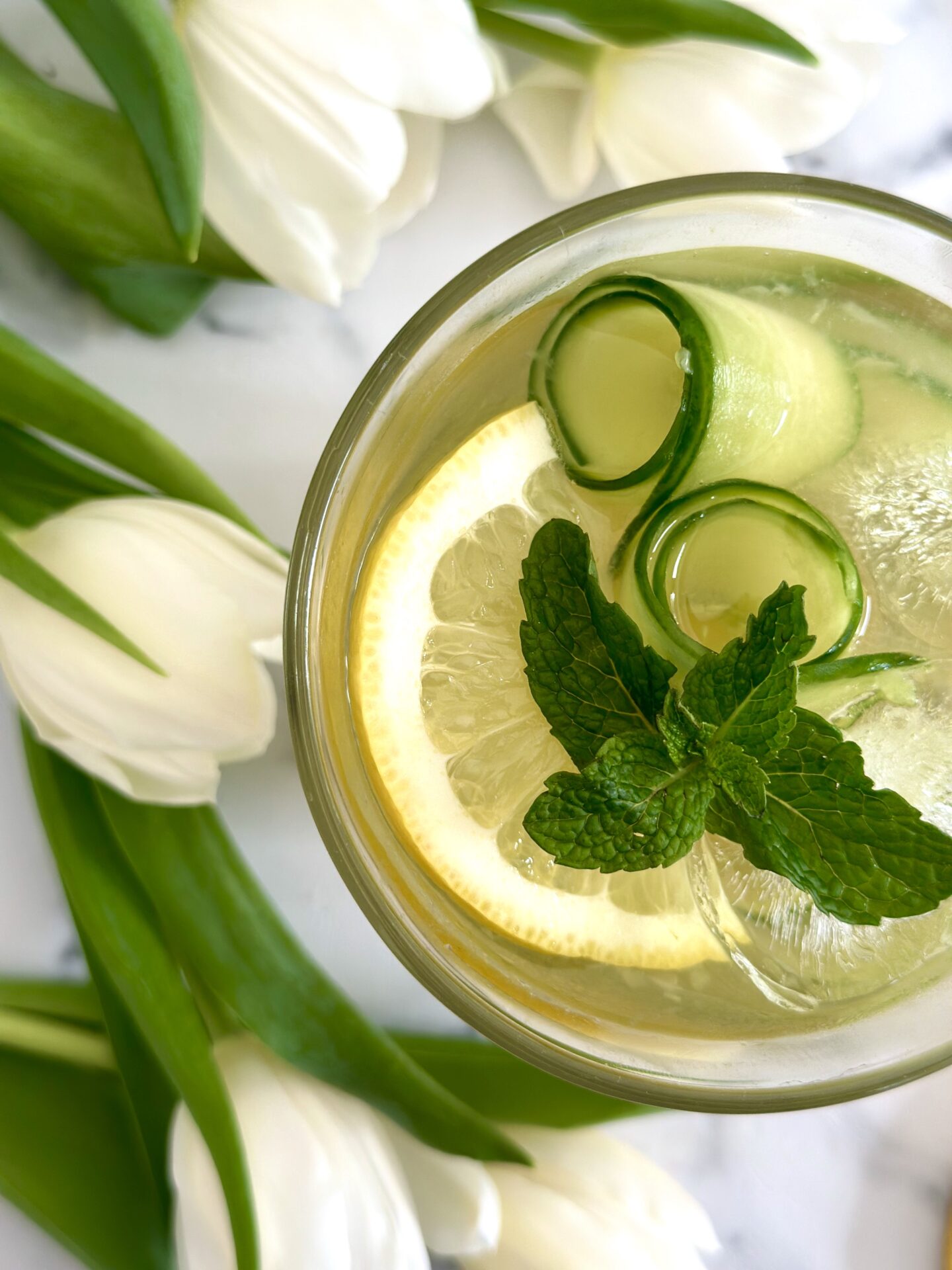 An Irish Maid Cocktail is seen from above garnished with cucumber curls, lemon slices and fresh mint and surrounded by white tulips