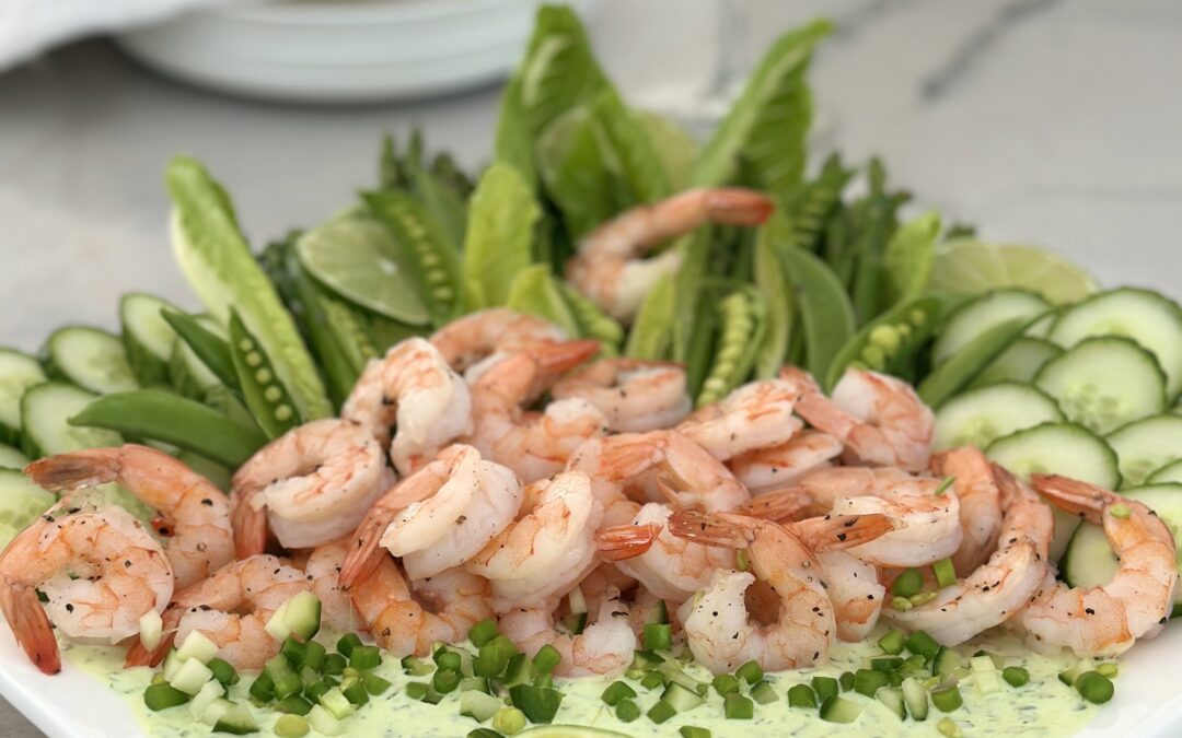A platter of shrimp with spring vegetables and creamy herb dip is seen on a white marble table with a glass of white wine