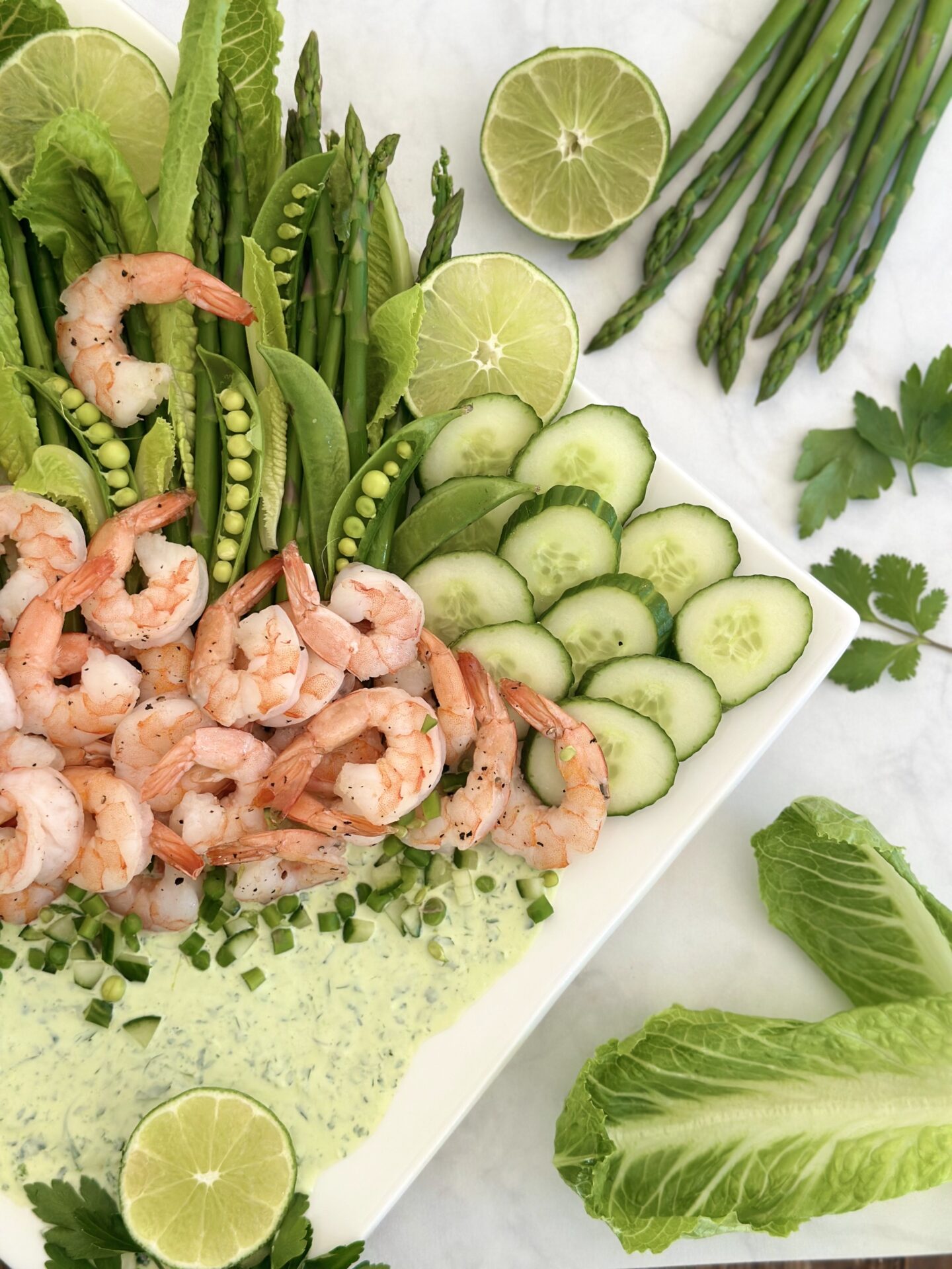 Half of a platter of shrimp with spring vegetables and a creamy herb dip is seen from above surrounded by fresh herb springs and fresh veggies