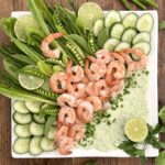 A beautiful platter of roasted shrimp with asparagus, cucumber, snap peas and romain is seen from above garnished with fresh herbs and lime slices