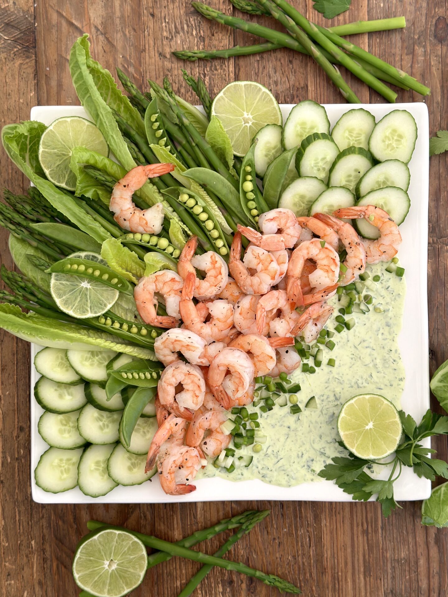 A beautiful platter of roasted shrimp with asparagus, cucumber, snap peas and romain is seen from above garnished with fresh herbs and lime slices