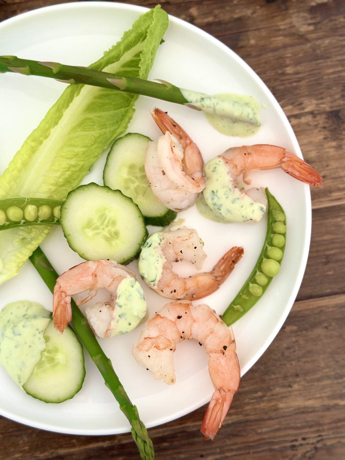 A white plate with a serving of roasted shrimp and spring vegetables with creamy herb dip is seen from above on a wood table
