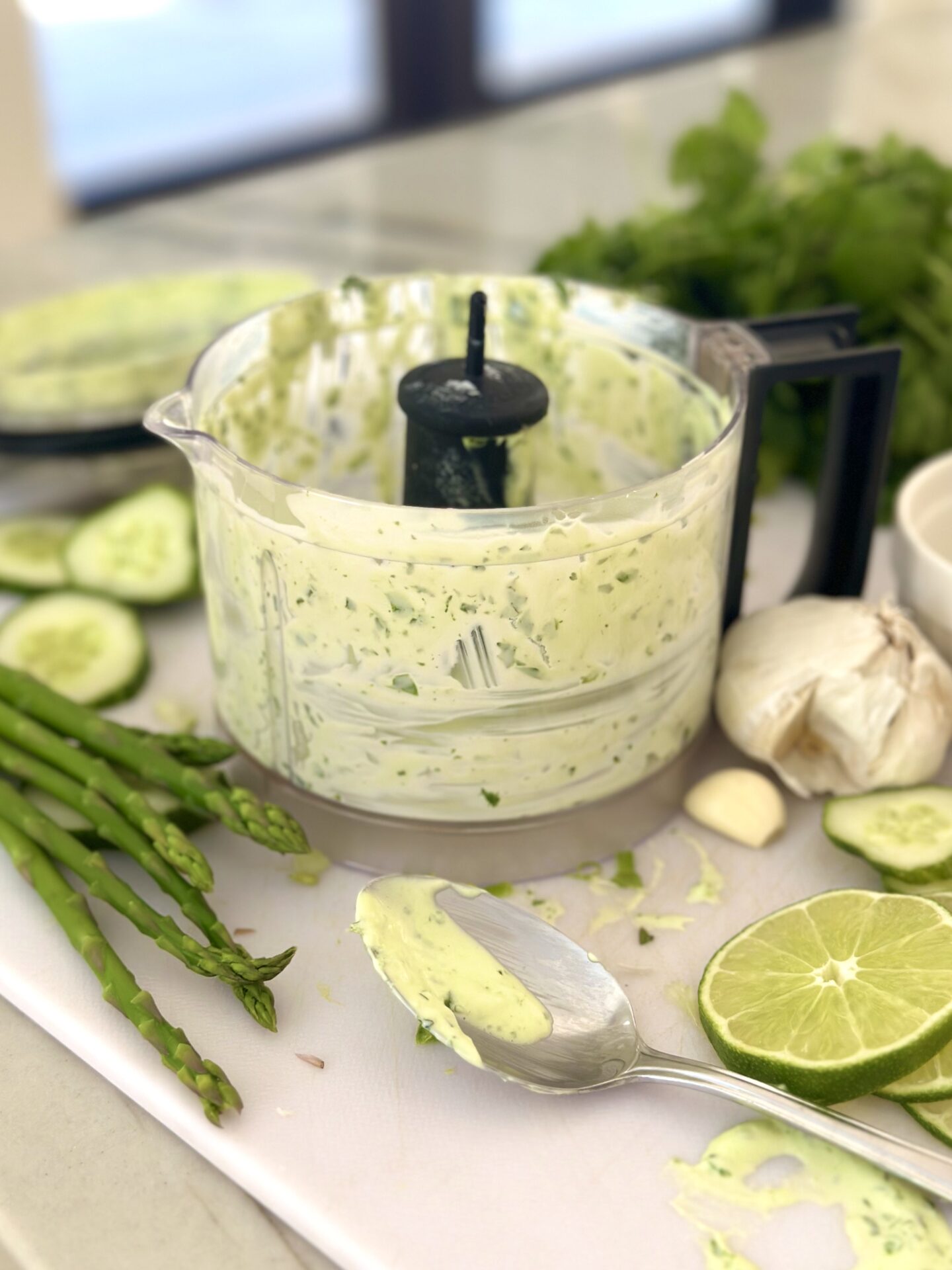 The bowl of a food processor is seen filled with creamy herb dip and surrounded by asparagus, lime, cucumber and fresh herbs