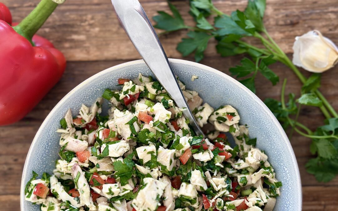 A bowl of chimichurri chicken salad is seen from above surrounded by a red bell pepper, garlic cloves and lots of fresh green herbs