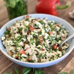 A bowl of chimichurri chicken salad is seen on a wood table surrounded by fresh parsley, cilantro, oregano, red bell pepper and garlic