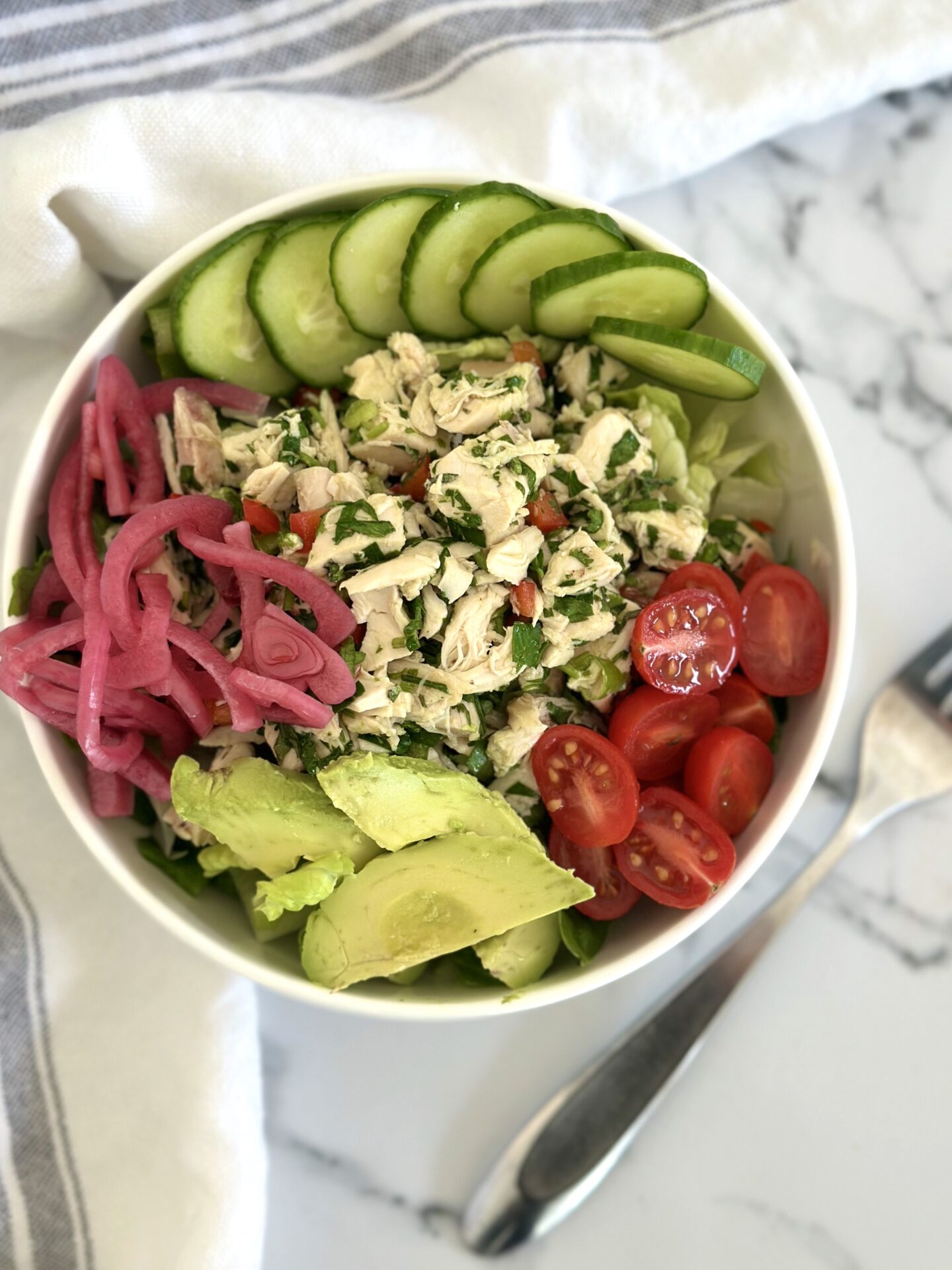 A green salad is topped with mounds of cherry tomatoes, avocado, cucumber, pickled red onion and a large scoop of chimichurri chicken salad
