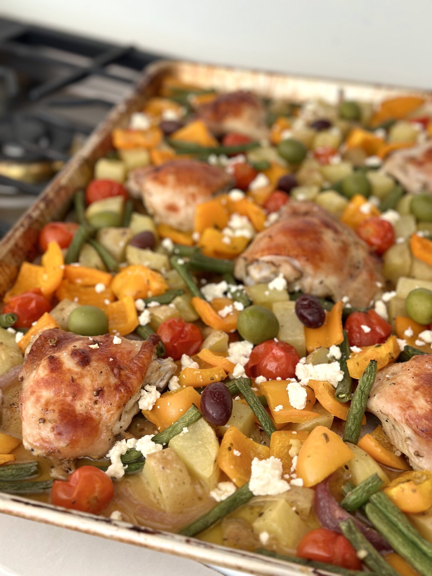 A large pan of Greek Sheet Pan Dinner that has roasted to perfection in the oven is seen resting on a cook top. The dinner features golden chicken thighs nestled amongst roasted orange bell peppers, chopped potatoes, red onion wedges and green beans. The meal is garnished with crumbled feta cheese and olives.