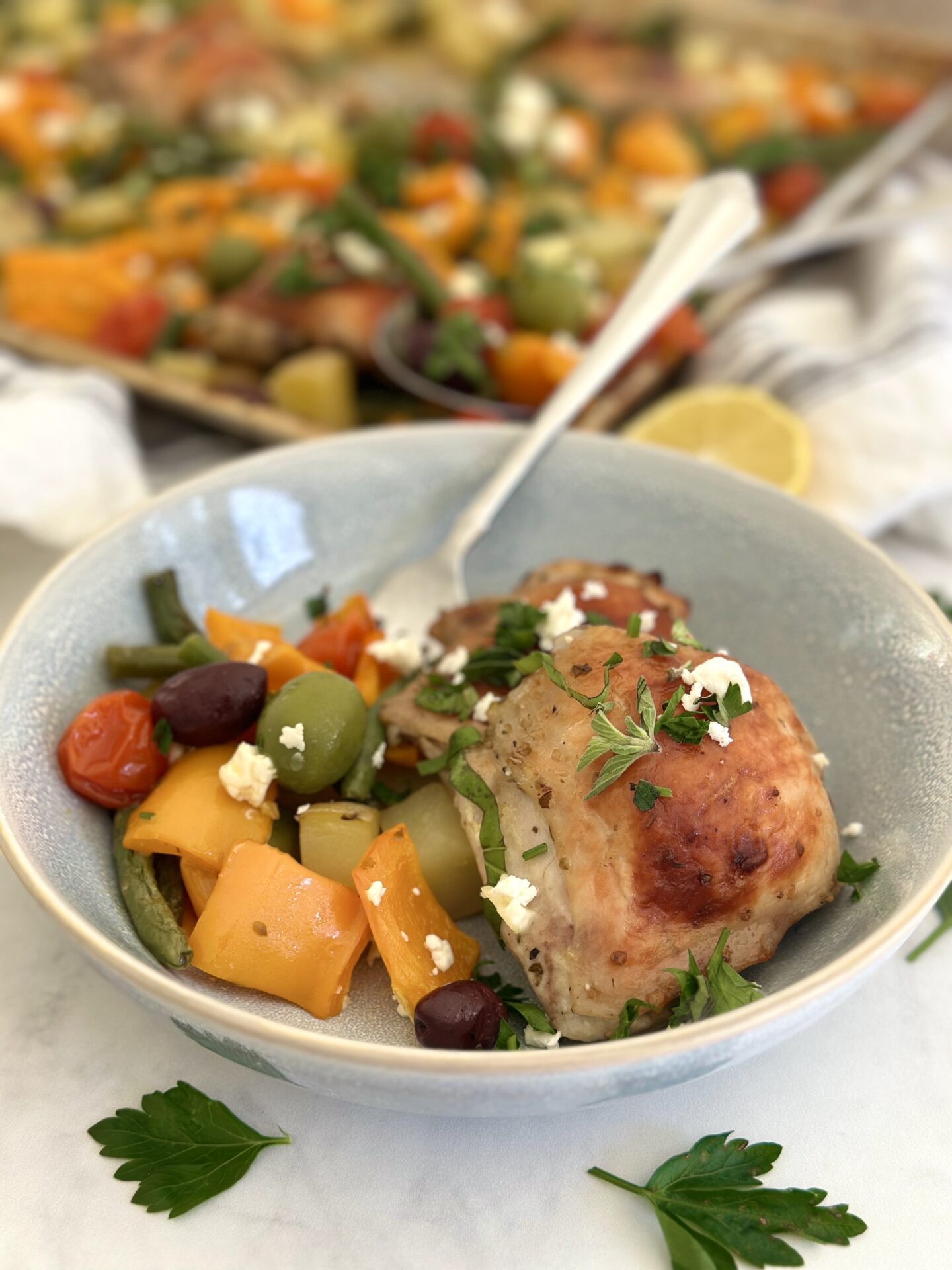 A bowl of Greek Chicken Sheet Pan Dinner is seen with golden chicken thighs, roasted vegetables, olives and feta cheese.  The sheet pan with the rest of the dinner is seen in the background.