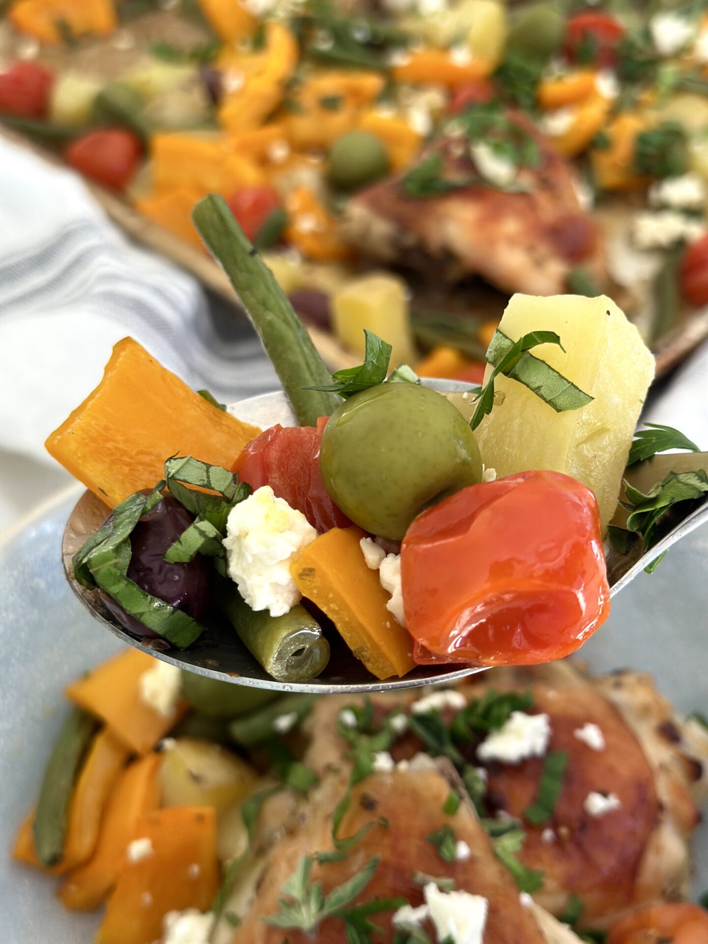 A large spoonful of vegetables, olives, feta cheese and fresh herbs is seen in the foreground with Greek Chicken Sheet Pan Dinner seen in behind