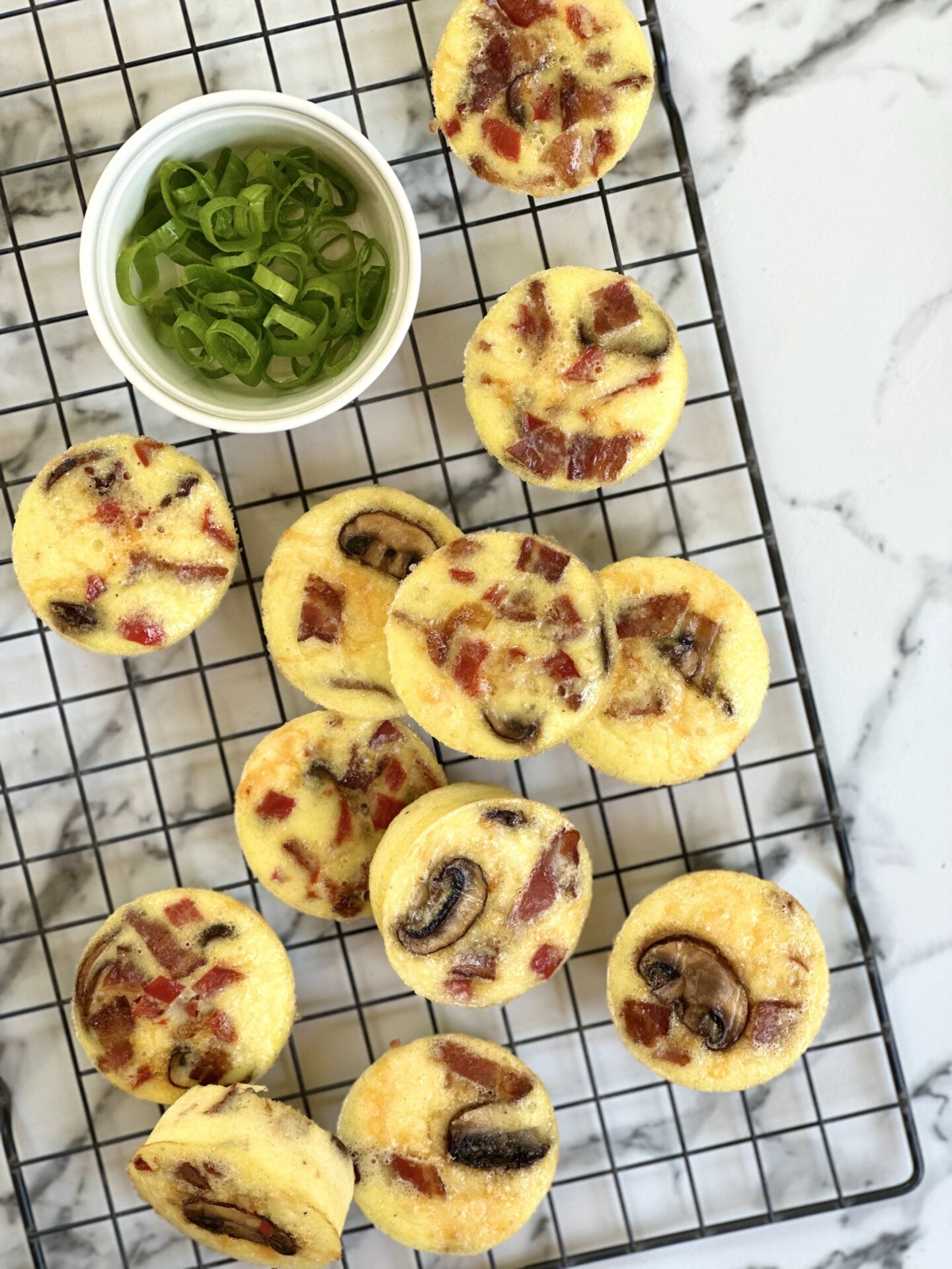 A cooling rack filled with oven baked egg bites is seen from above with a dish of chopped green onions