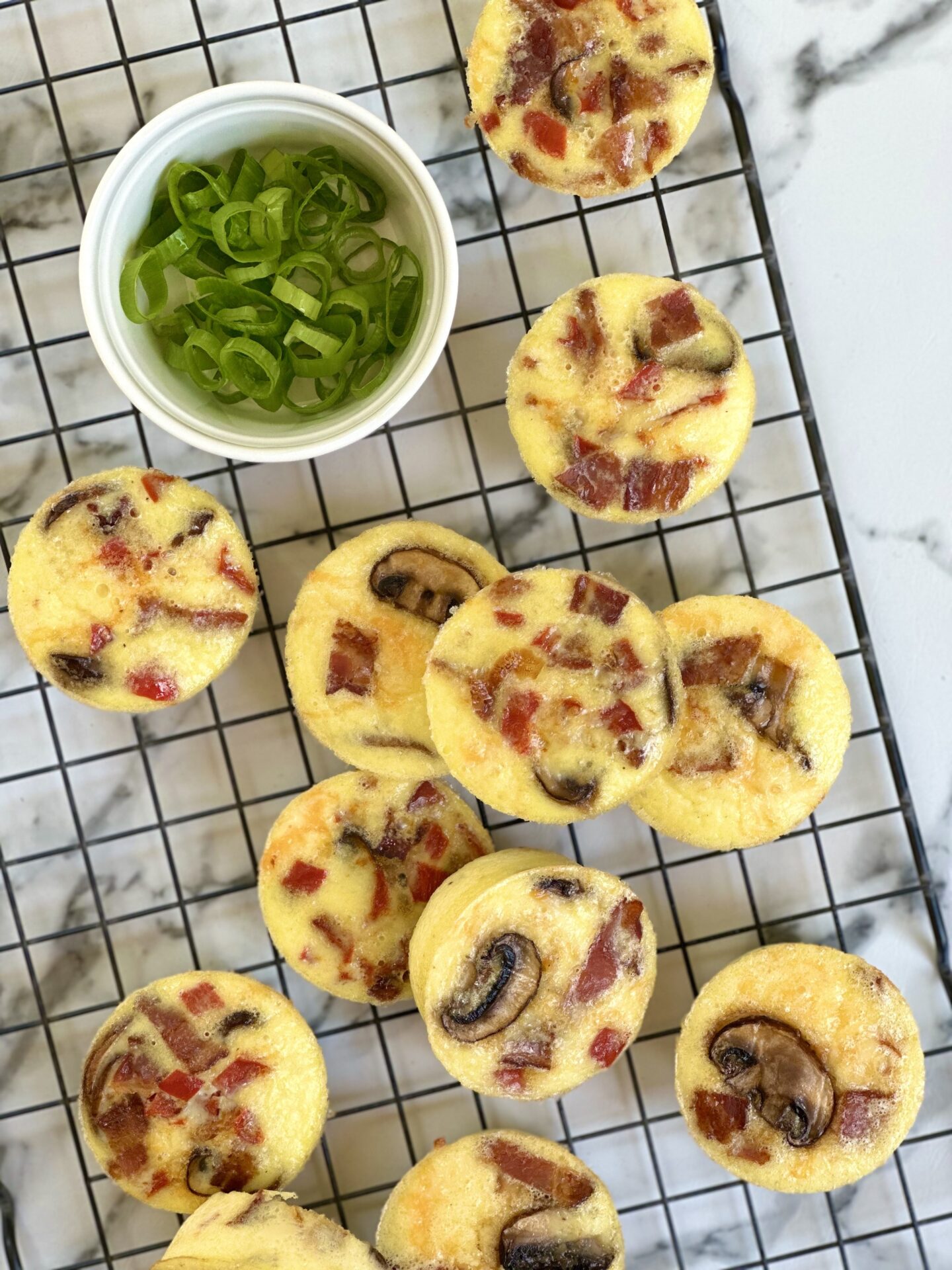 A batch of oven baked egg bites with bacon, mushroom and peppers is seen from above, cooling on a black wire rack