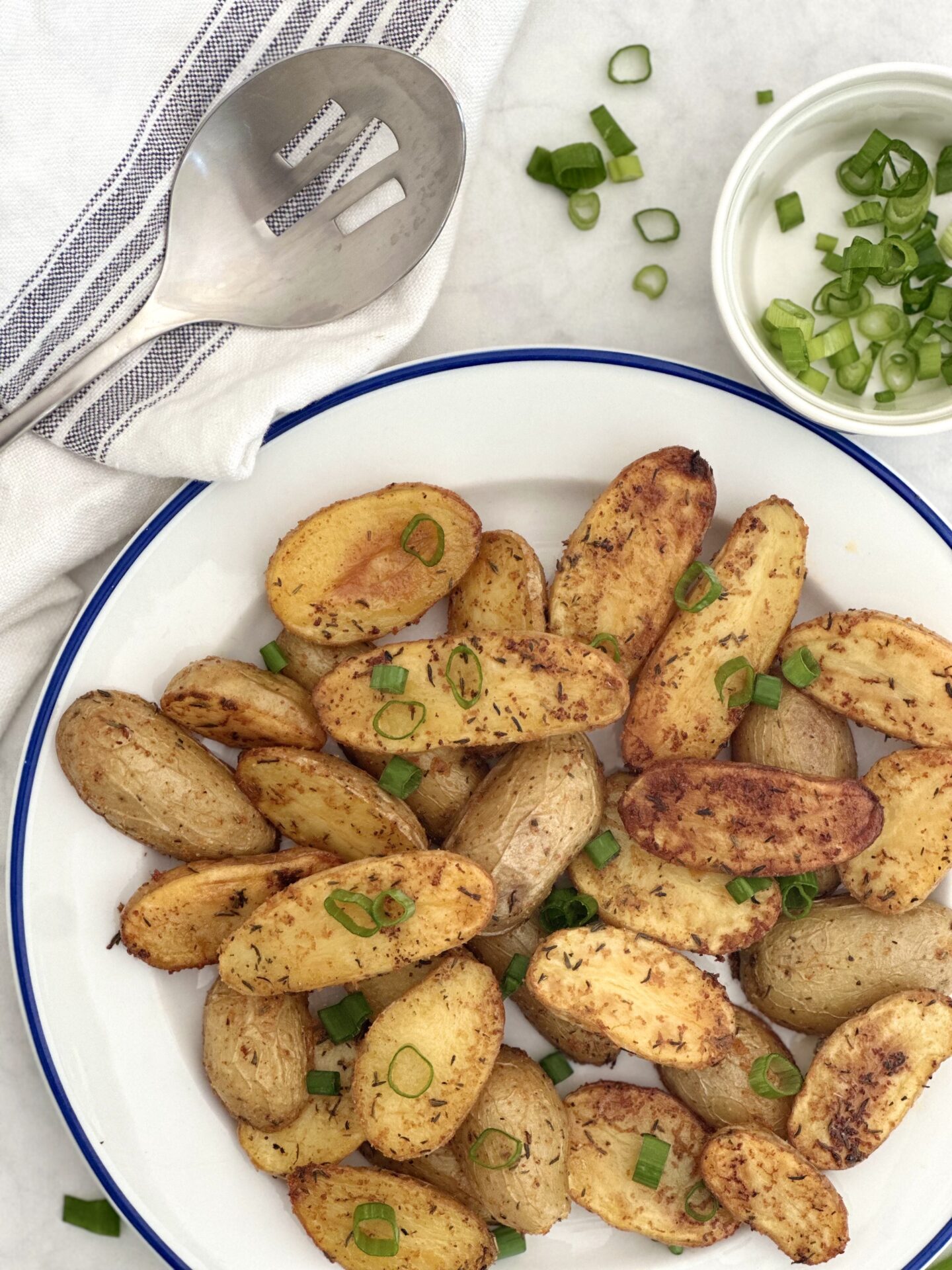 A blue trimmed plate of golden and crispy roasted fingerling potatoes is seen from above with a serving spoon and chopped green onion for garnishing