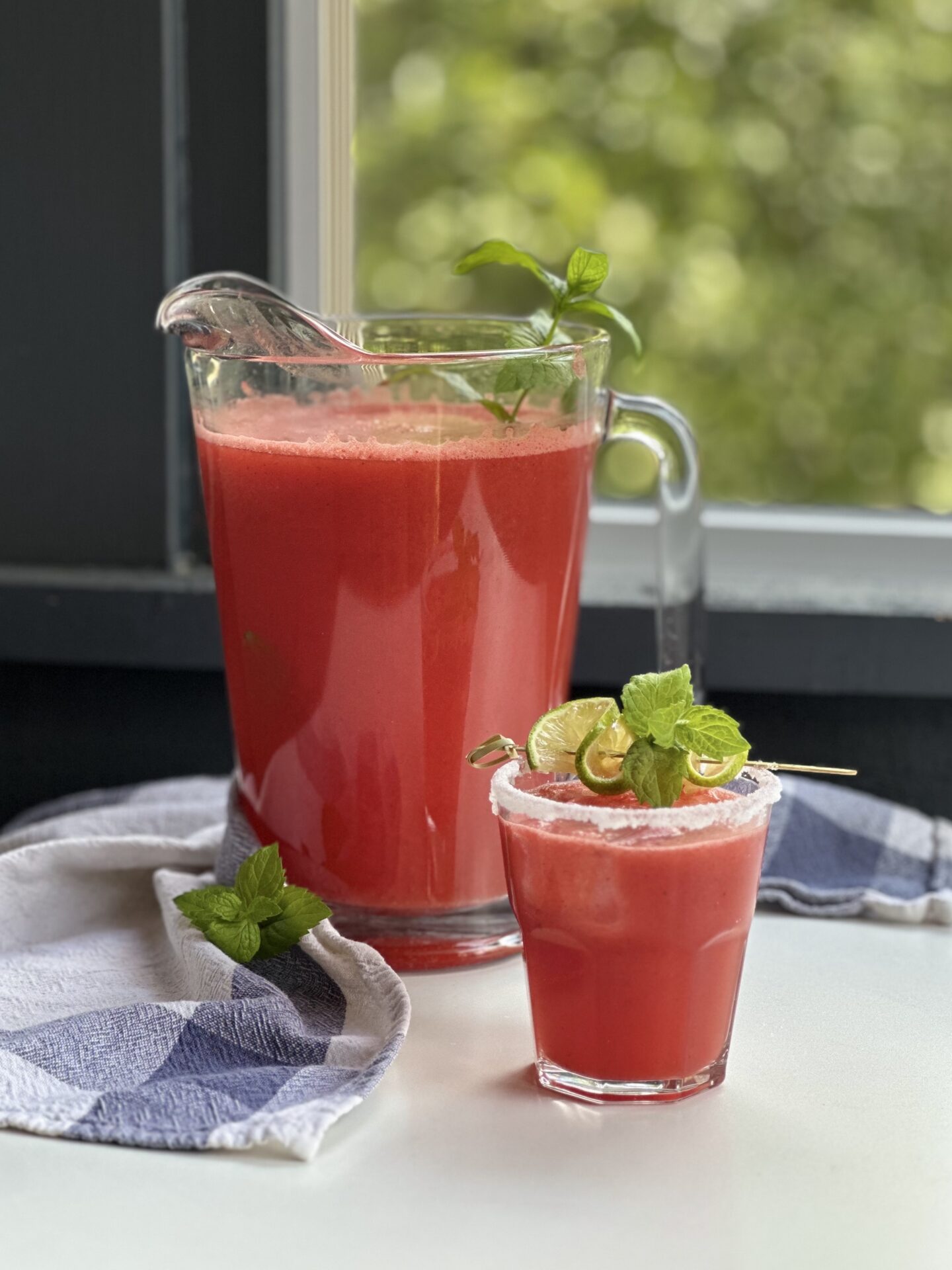 A large pitcher of freshly made watermelon agua fresca sits by a window, with a sugar rimmed glass of the beverage, garnished with lime and mint, sits in the foreground.  